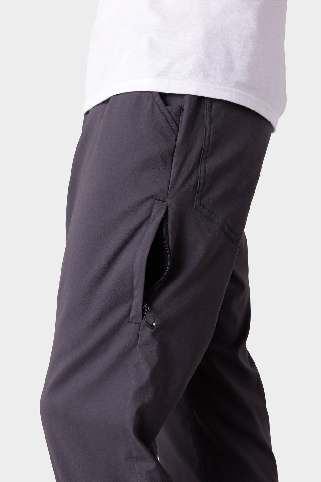 A person models the 686 Men's Everywhere® Merino-Lined Pant - Relaxed Fit in dark gray, featuring a zipper pocket, paired with a white shirt and sleek insulated outerwear, shown from waist to mid-thigh against a plain light background.