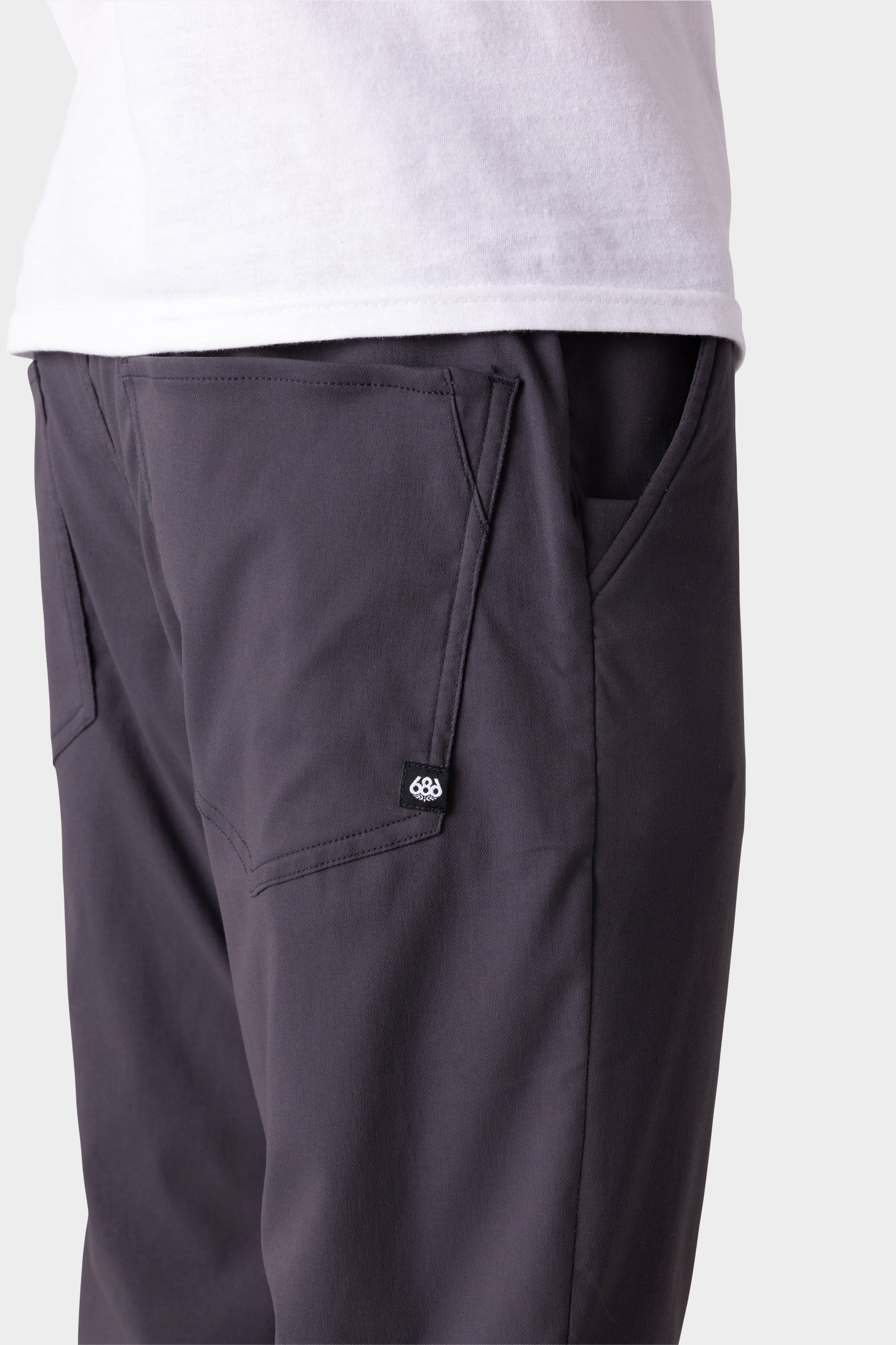 A person wears 686 Men's Everywhere® Merino-Lined Pant - Relaxed Fit in dark gray, featuring visible front pockets and a small logo patch, paired with a white shirt and outerwear. The image highlights the pants' pocket details from the waist down.
