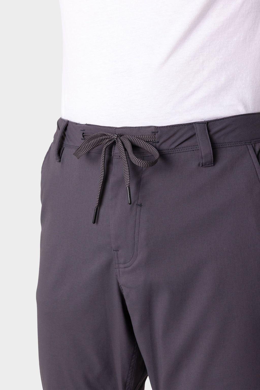 A person wears 686 Men's Everywhere® Merino-Lined Pant - Relaxed Fit in dark gray, featuring a drawstring waist tied in a bow, paired with a white shirt and stylish insulated outerwear. The photo shows the pants from waist to mid-thigh.