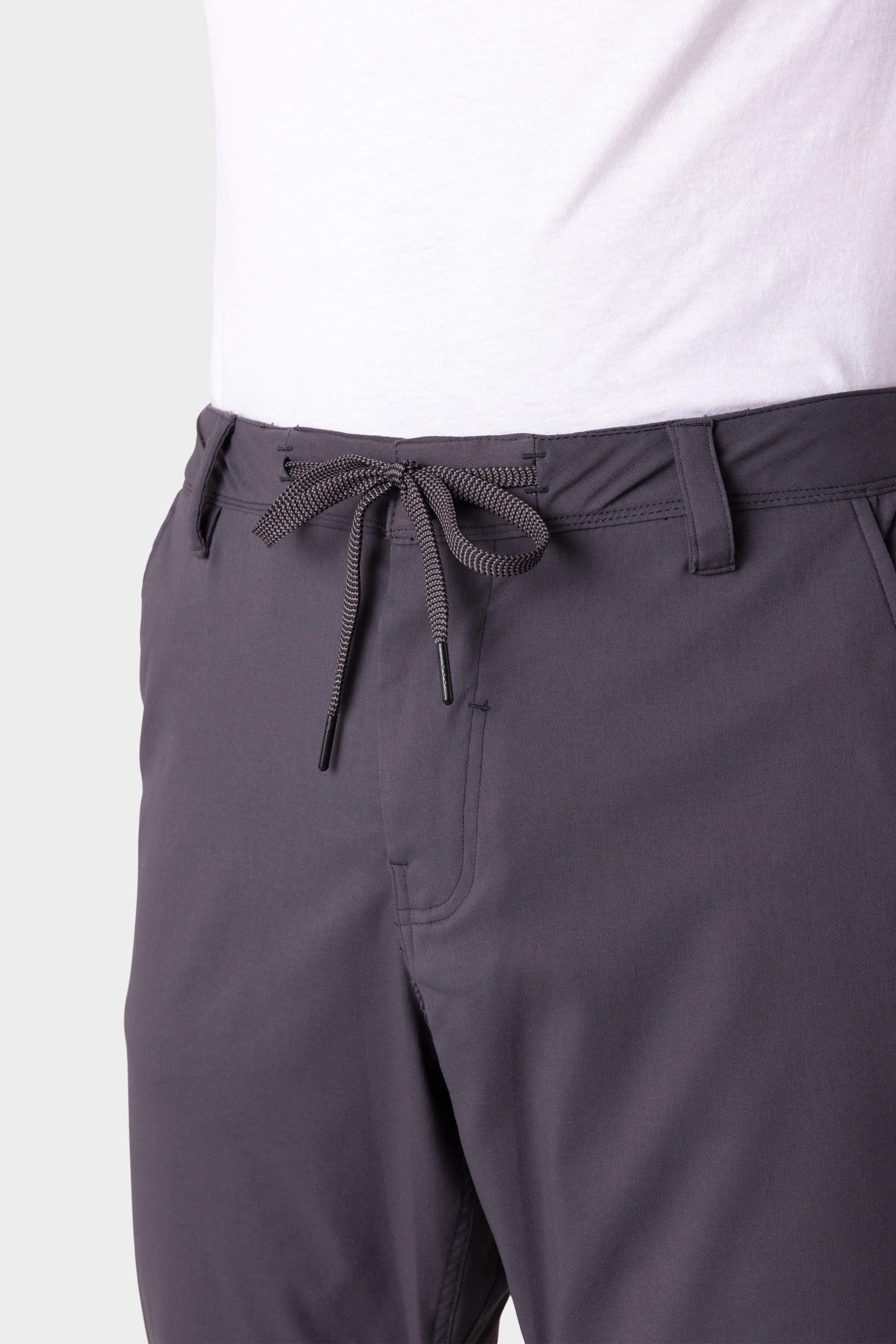 A person wears 686 Men's Everywhere® Merino-Lined Pant - Relaxed Fit in dark gray, featuring a drawstring waist tied in a bow, paired with a white shirt and stylish insulated outerwear. The photo shows the pants from waist to mid-thigh.