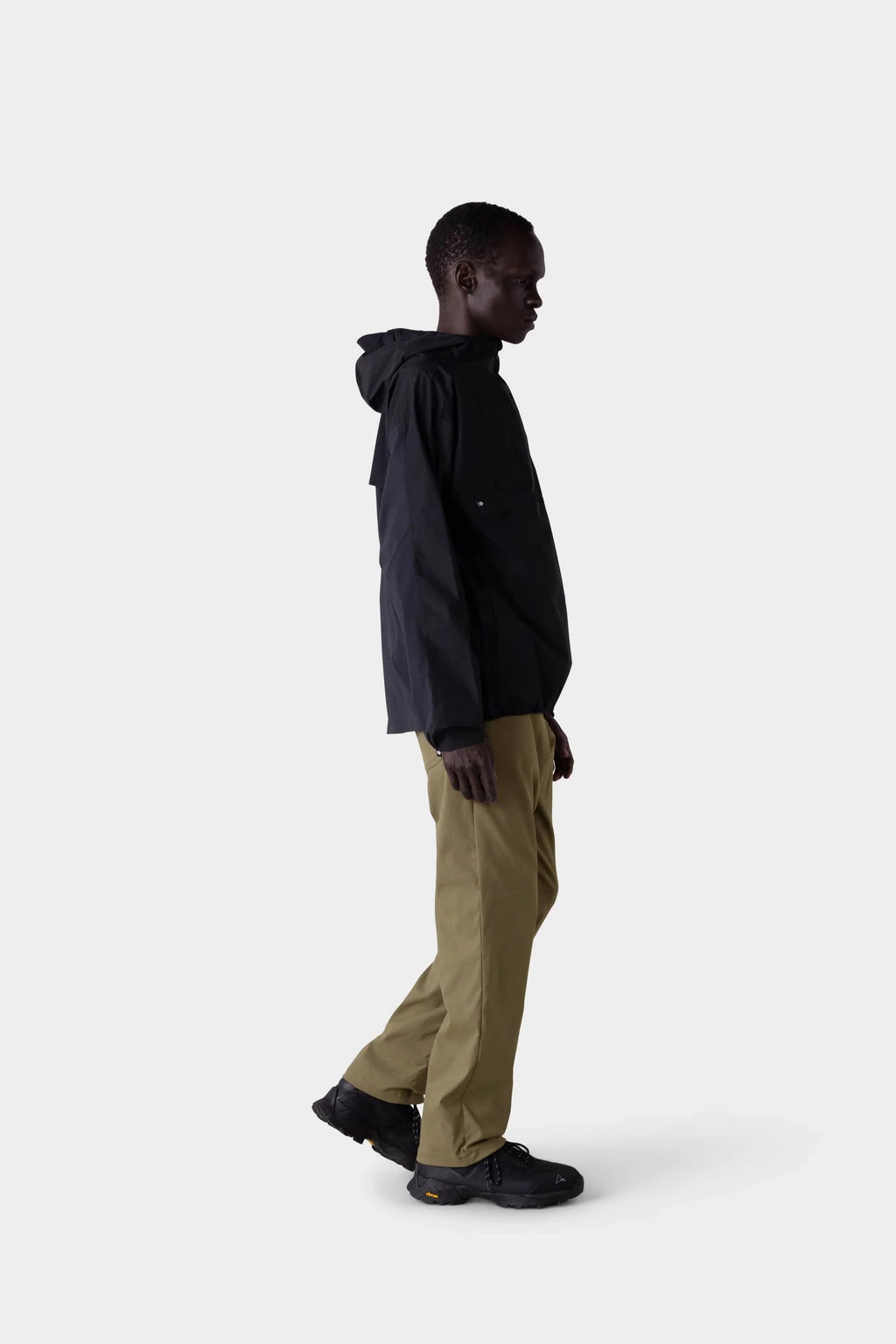 A person stands in profile against a plain light background, wearing a stylish black hooded jacket, black shoes, and 686 Men's Everywhere® Merino-Lined Pant - Relaxed Fit by 686.