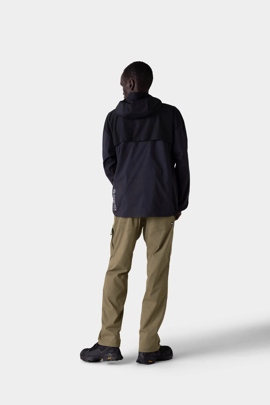 A person stands facing away, wearing a stylish insulated black hooded winter jacket, 686 Men's Everywhere® Merino-Lined Pant - Relaxed Fit by 686 in khaki, and black shoes against a plain light gray background.