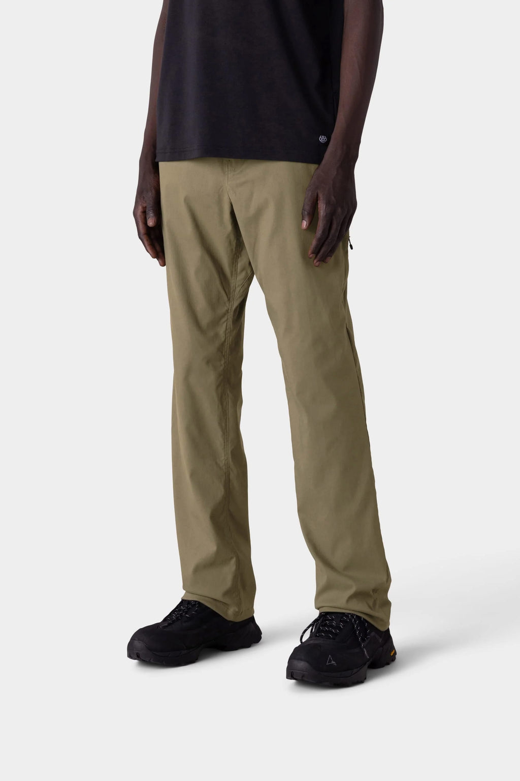 The lower half of a person is shown against a light gray background, wearing the 686 Men's Everywhere® Merino-Lined Pant - Relaxed Fit in olive green, paired with a black shirt, black shoes, and sleek insulated outerwear.