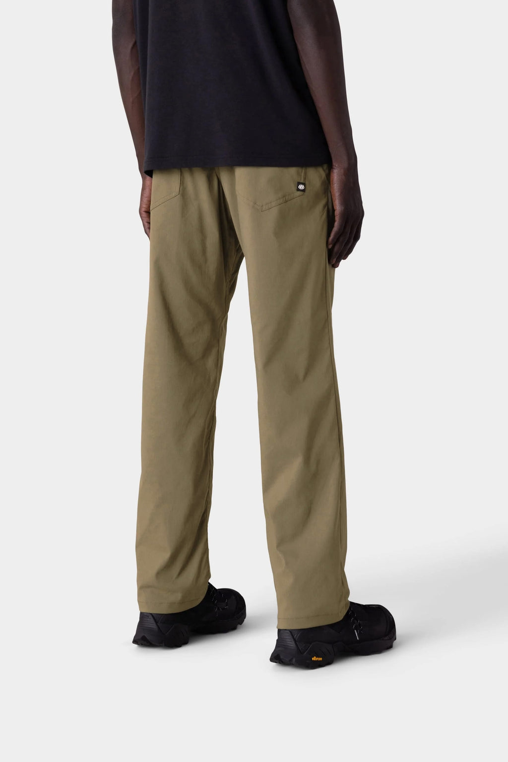 A person stands facing away in 686 Men's Everywhere® Merino-Lined Pant - Relaxed Fit, paired with a black shirt and black shoes, styled with insulated outerwear against a plain light gray background.
