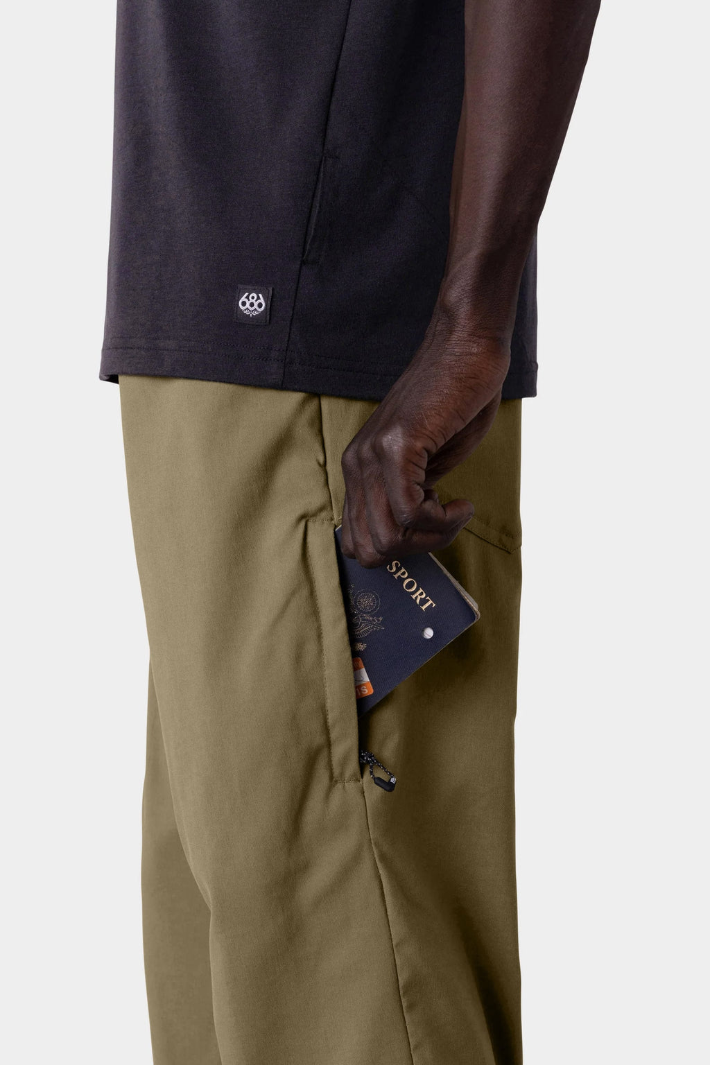 Wearing a 686 Men's Everywhere® Merino-Lined Pant - Relaxed Fit in olive, a person puts a navy passport and boarding pass into the zippered side pocket, pairing the pants with a dark shirt and warm winter jacket.