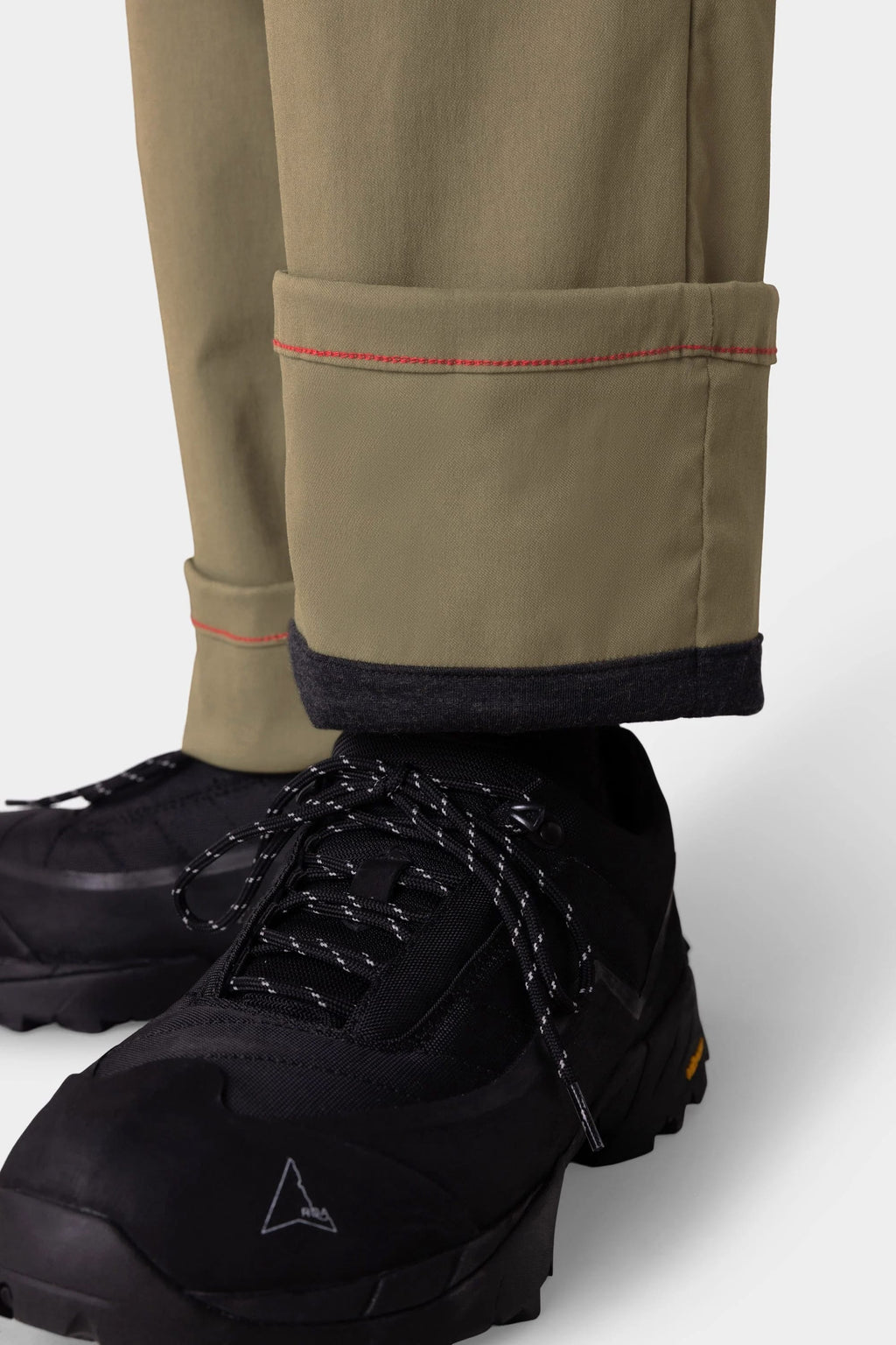 A close-up shows someone in 686 Men's Everywhere® Merino-Lined Pant - Relaxed Fit with a folded cuff and red stitching, styled with black hiking shoes with white-speckled laces and a sleek insulated jacket against a plain light background.