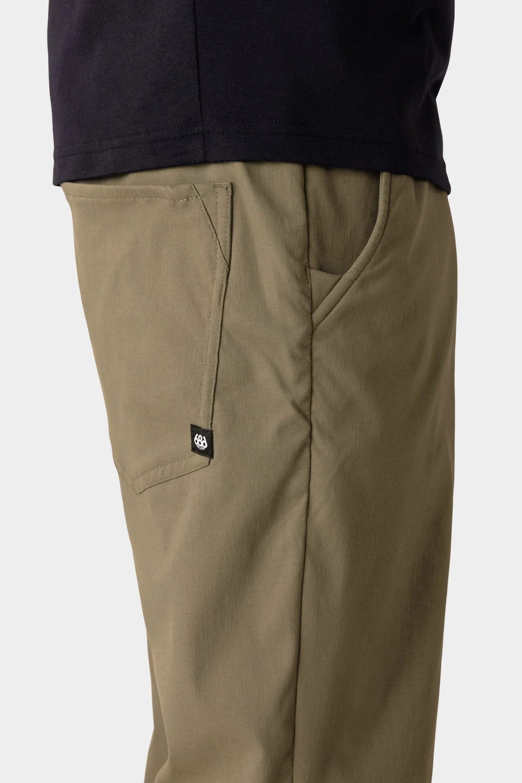 A person wearing 686 Men's Everywhere® Merino-Lined Pant - Relaxed Fit in olive with a slanted front pocket and black logo tag, paired with a black shirt and insulated outerwear. Image is cropped to show only the waist and upper thigh.