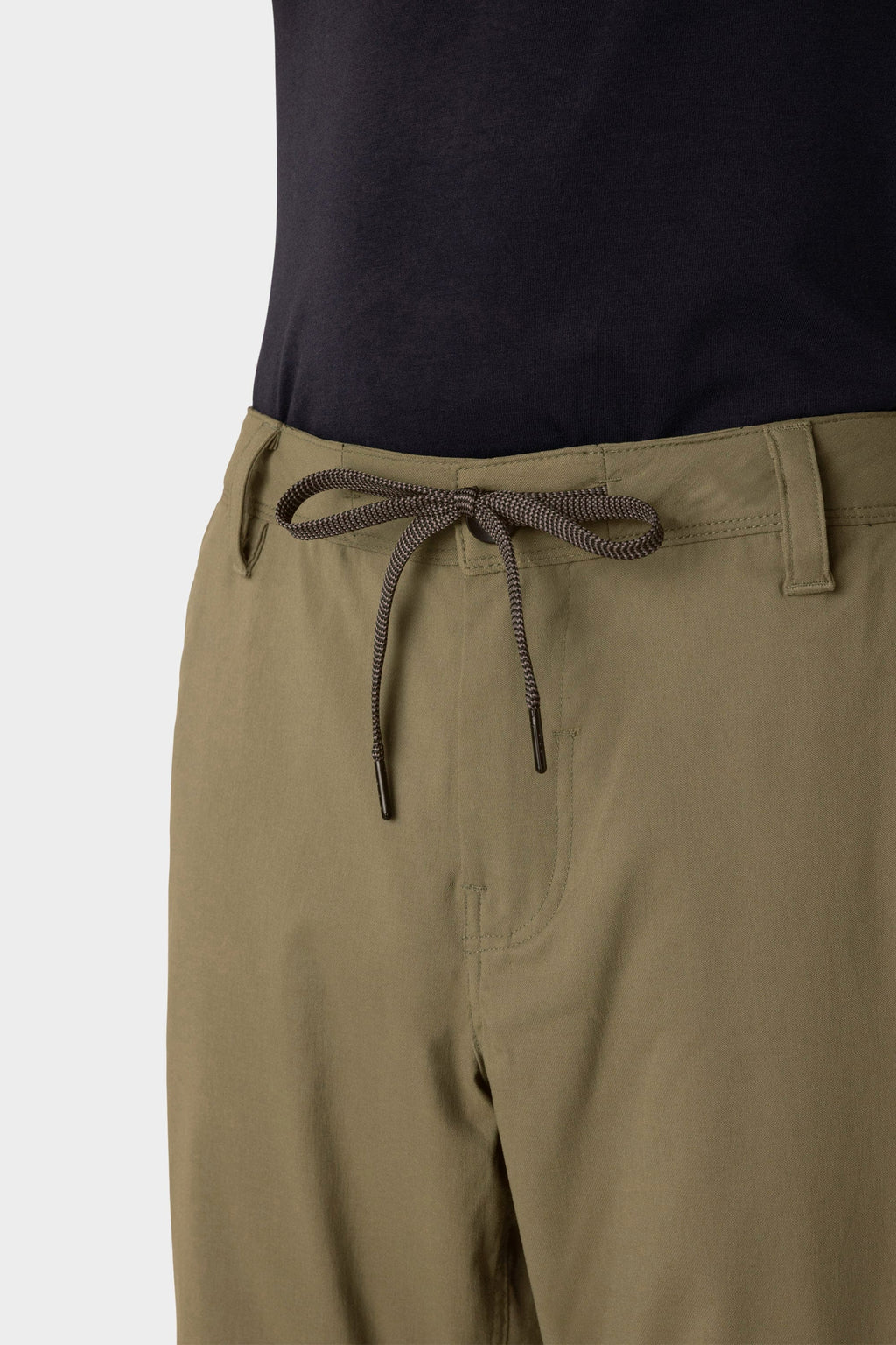 A close-up of a person in 686 Men's Everywhere® Merino-Lined Pant - Relaxed Fit in light olive, featuring a drawstring waist, paired with a tucked black shirt and a fashionable insulated outerwear layer for chilly days.