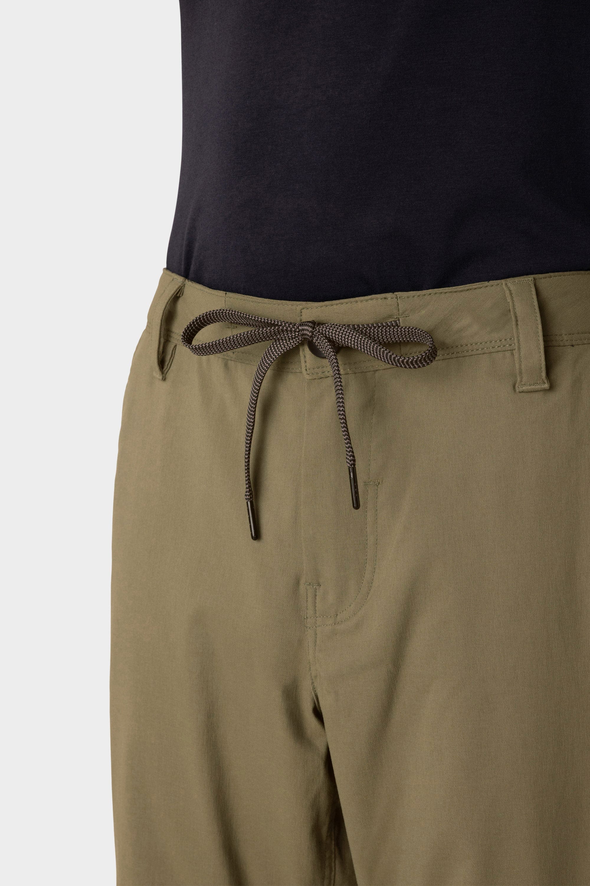 A close-up of a person in 686 Men's Everywhere® Merino-Lined Pant - Relaxed Fit in light olive, featuring a drawstring waist, paired with a tucked black shirt and a fashionable insulated outerwear layer for chilly days.