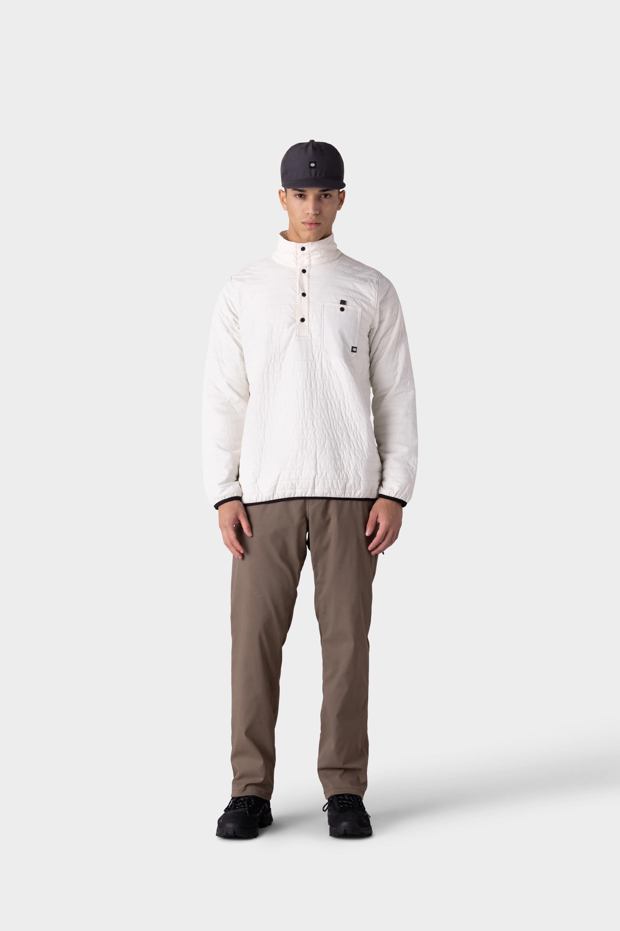 A person stands facing forward wearing a white quilted buttoned pullover, black shoes, a dark gray cap, and 686 Men's Everywhere® Merino-Lined Pant - Relaxed Fit by 686, against a plain light gray background.