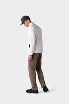 A man wearing a gray cap, stylish insulated jacket, and black shoes models the 686 Men's Everywhere® Merino-Lined Pant - Relaxed Fit in brown, standing slightly turned against a plain light gray background.
