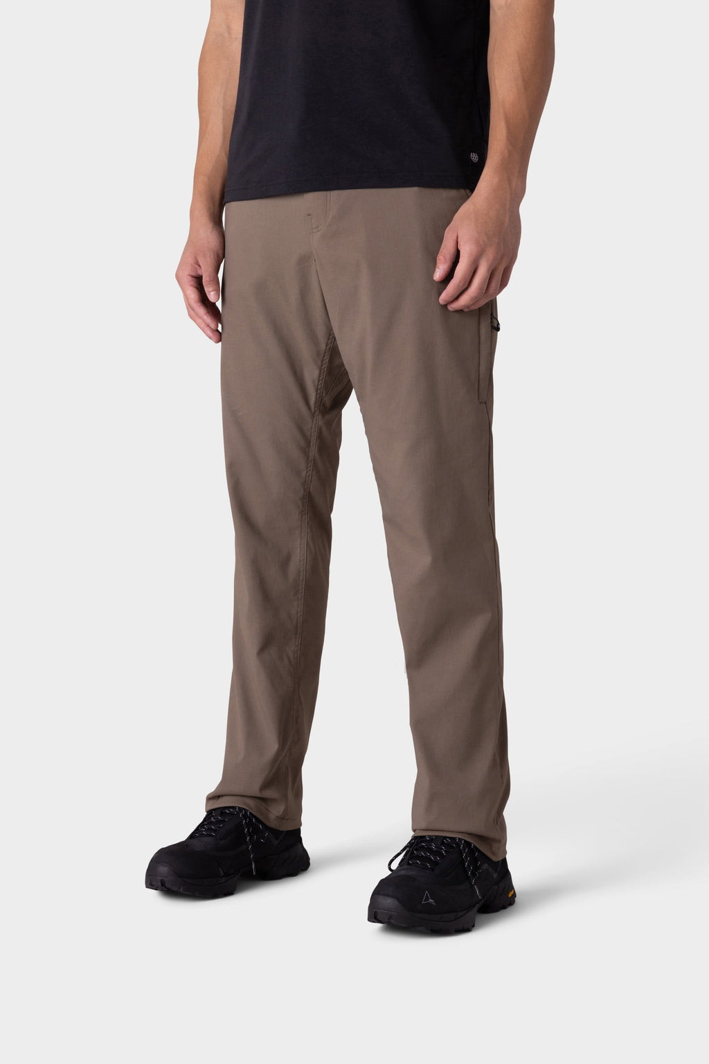 A person is wearing 686 Men's Everywhere® Merino-Lined Pant - Relaxed Fit in brown, paired with black shoes, standing against a plain white background. The image is cropped at the shoulders, showing only the lower half of the body.