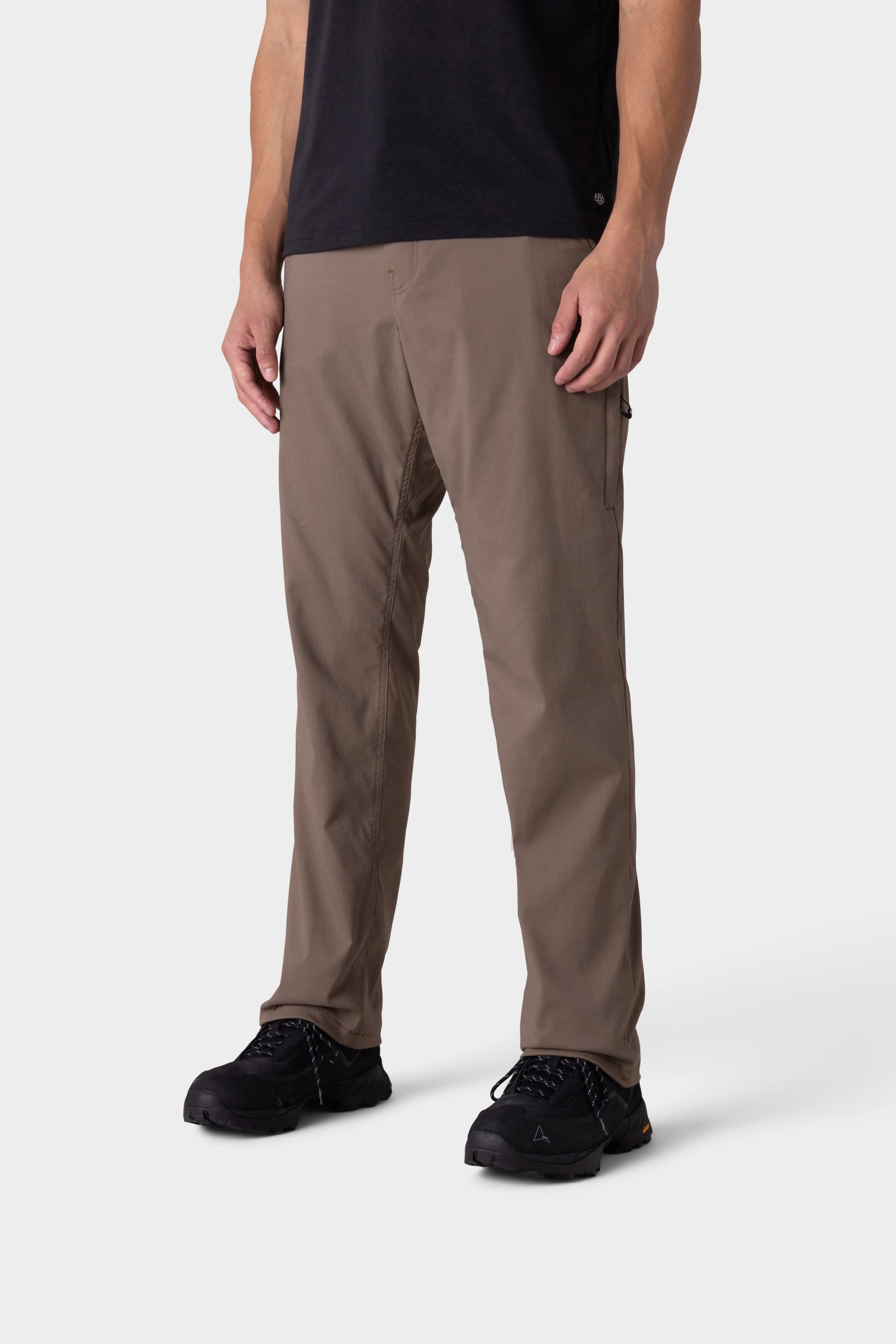 A person is wearing 686 Men's Everywhere® Merino-Lined Pant - Relaxed Fit in brown, paired with black shoes, standing against a plain white background. The image is cropped at the shoulders, showing only the lower half of the body.