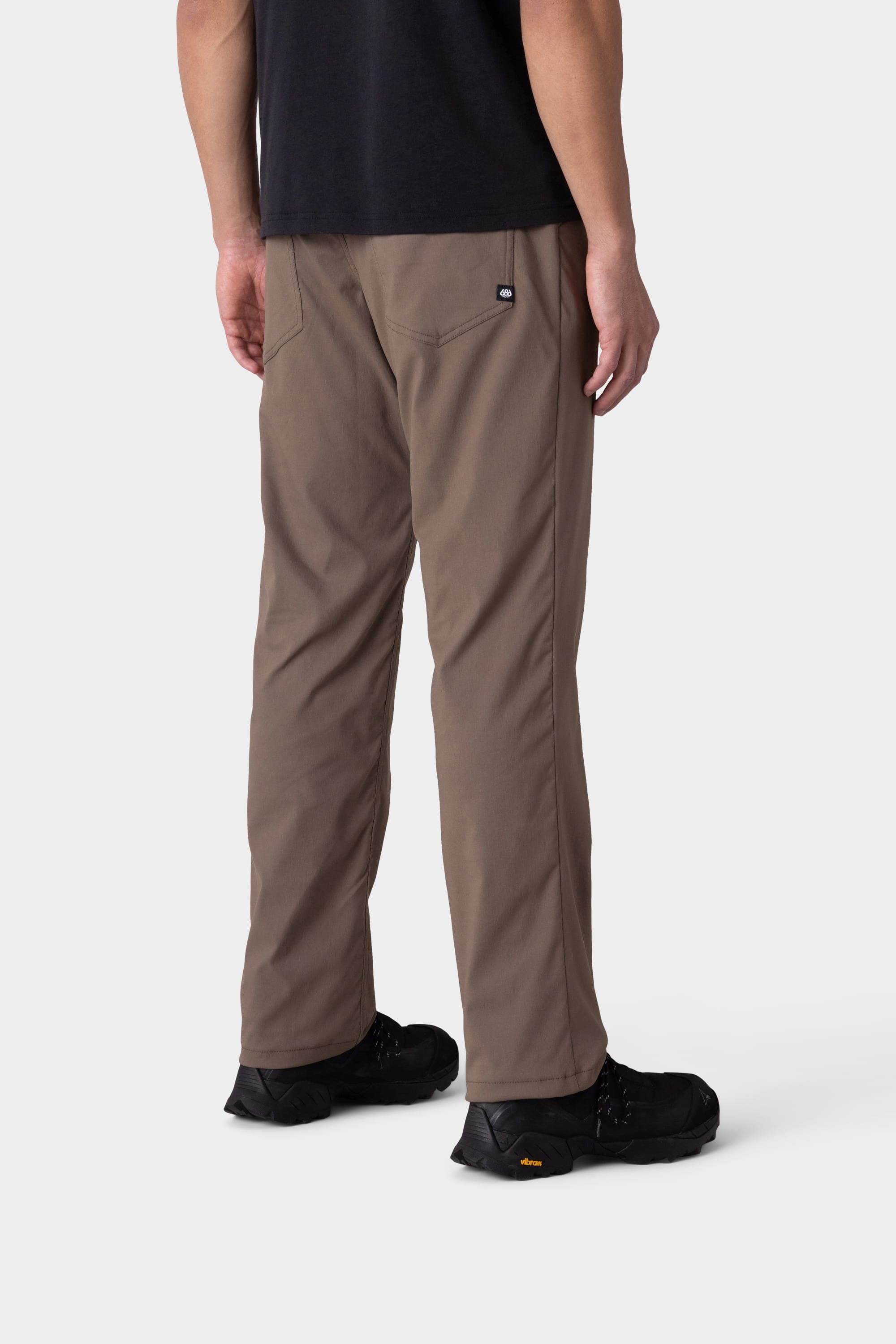 A person wearing stylish insulated outerwear, 686 Men's Everywhere® Merino-Lined Pant - Relaxed Fit by 686 in light brown, a black t-shirt, and black shoes stands facing away from the camera against a plain light background.