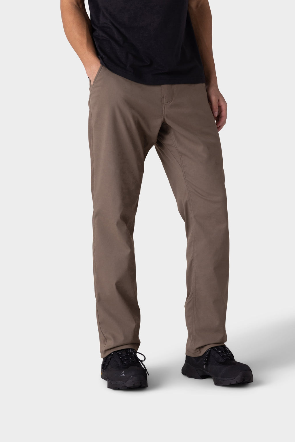 A person wears 686 Men's Everywhere® Merino-Lined Pant - Relaxed Fit by 686, a black t-shirt, a warm winter jacket, and black shoes, standing with hands in pockets against a plain light gray background.