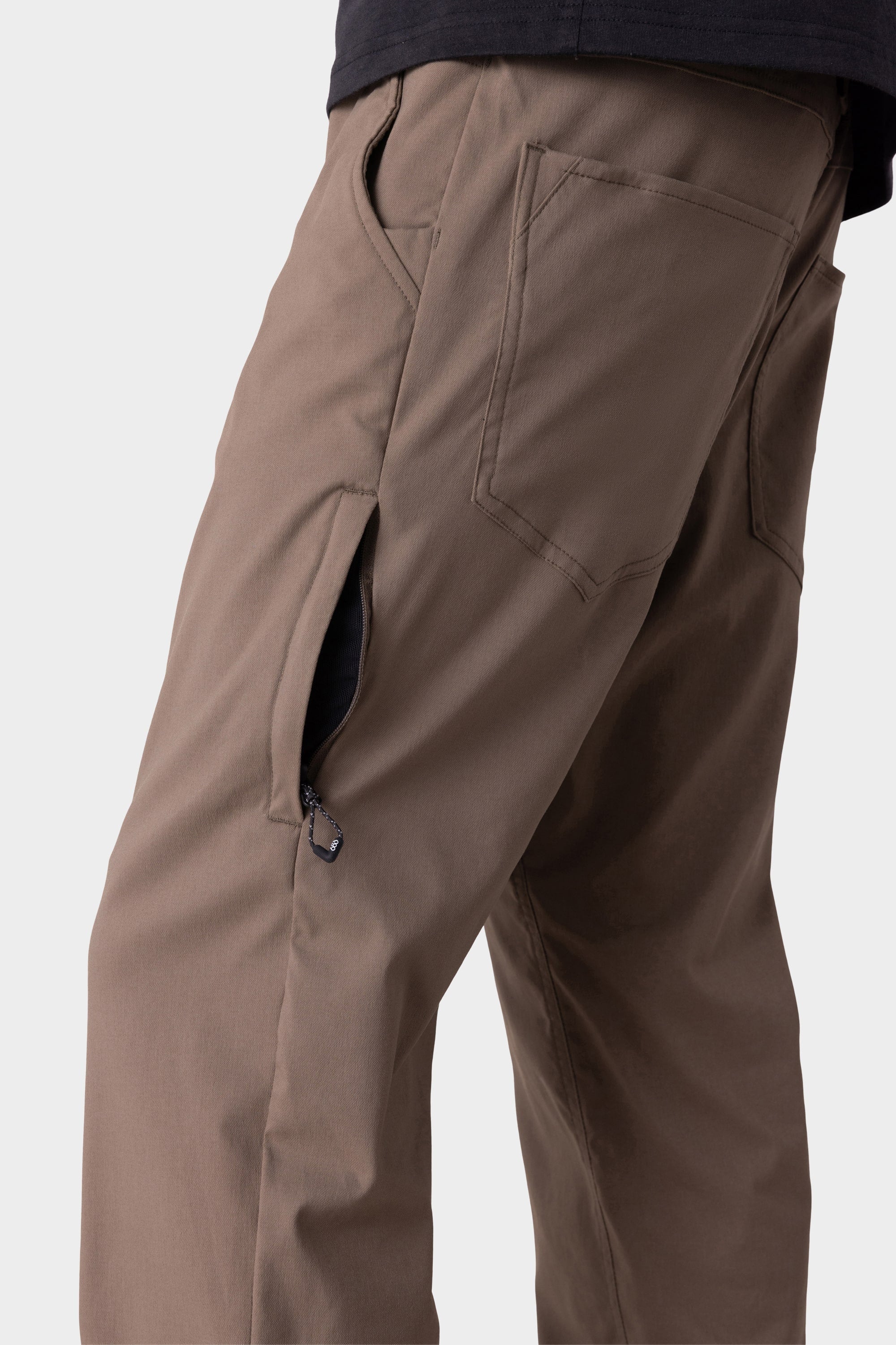 A close-up of a person wearing 686 Men's Everywhere® Merino-Lined Pant - Relaxed Fit in brown, featuring visible front and back pockets, a partially open side zippered pocket, paired with a black top and stylish insulated outerwear for chilly days.
