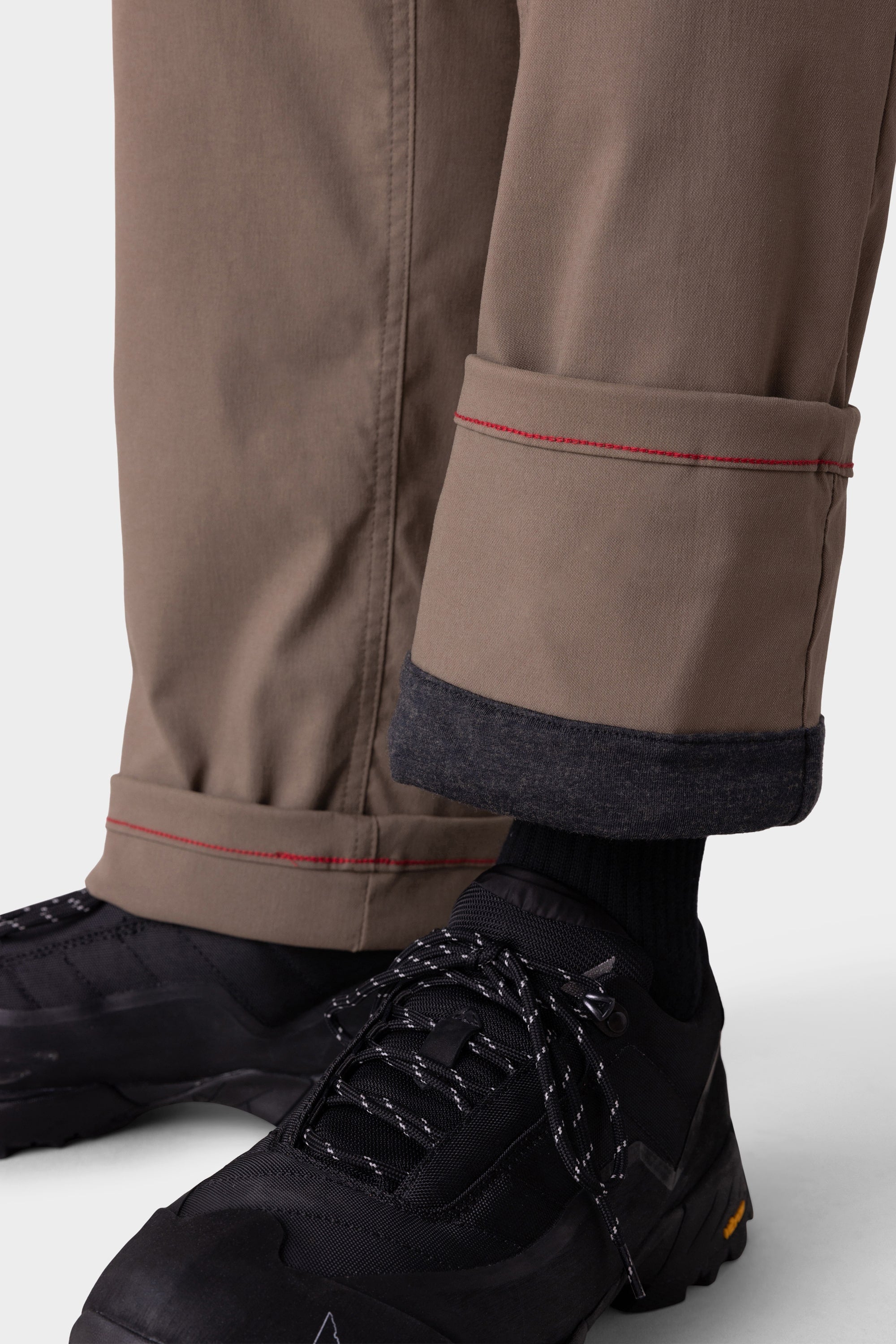 A close-up of a person wearing 686 Men's Everywhere® Merino-Lined Pant - Relaxed Fit in taupe with red stitching, revealing a dark gray inner lining, paired with black socks, black lace-up sneakers, and a stylish insulated outerwear for chilly days.