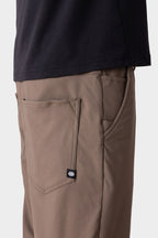 A close-up side view of a person wearing 686 Men's Everywhere® Merino-Lined Pant - Relaxed Fit in taupe with a back pocket and black logo tag, paired with a black shirt and insulated outerwear against a plain light background.