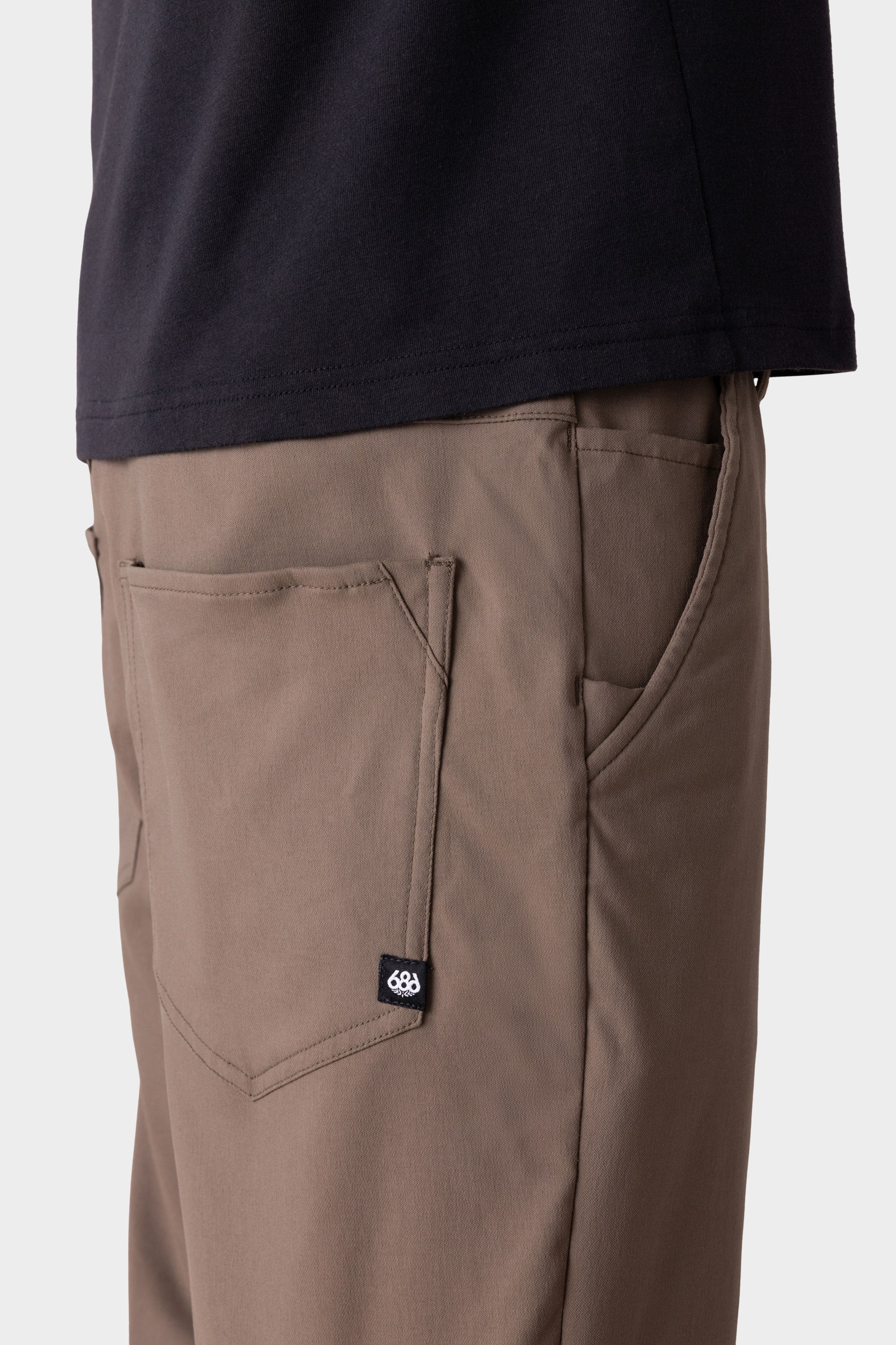 A close-up side view of a person wearing 686 Men's Everywhere® Merino-Lined Pant - Relaxed Fit in taupe with a back pocket and black logo tag, paired with a black shirt and insulated outerwear against a plain light background.