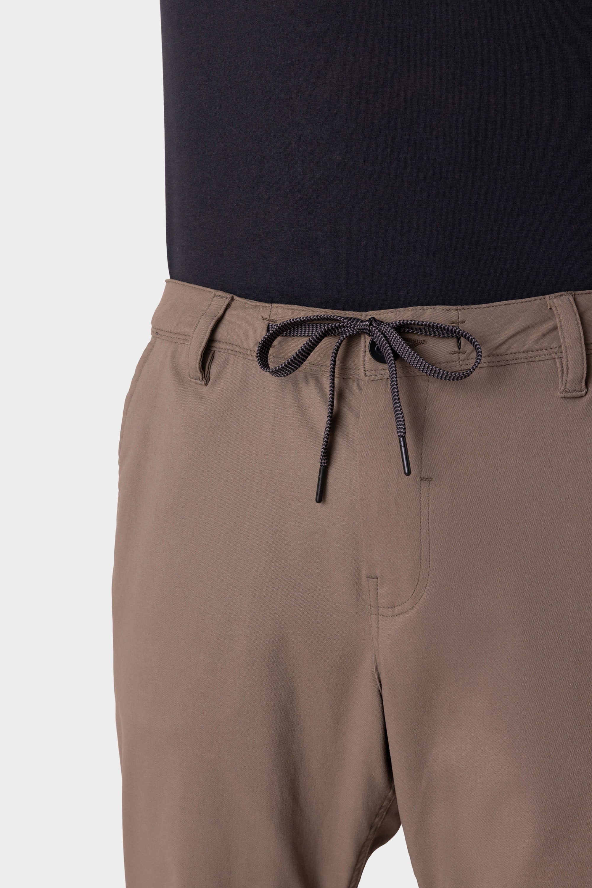 A close-up of a person wearing the 686 Men's Everywhere® Merino-Lined Pant - Relaxed Fit in light brown, paired with a black shirt and insulated outerwear, highlights the waistband, drawstring tie, and upper part of these 686 pants.