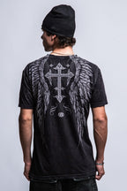 A man in a black beanie and the 686 Affliction Death Eyes SS Tee by 686, featuring large angel wings and a decorative cross graphic on the back, stands against a plain light gray background.
