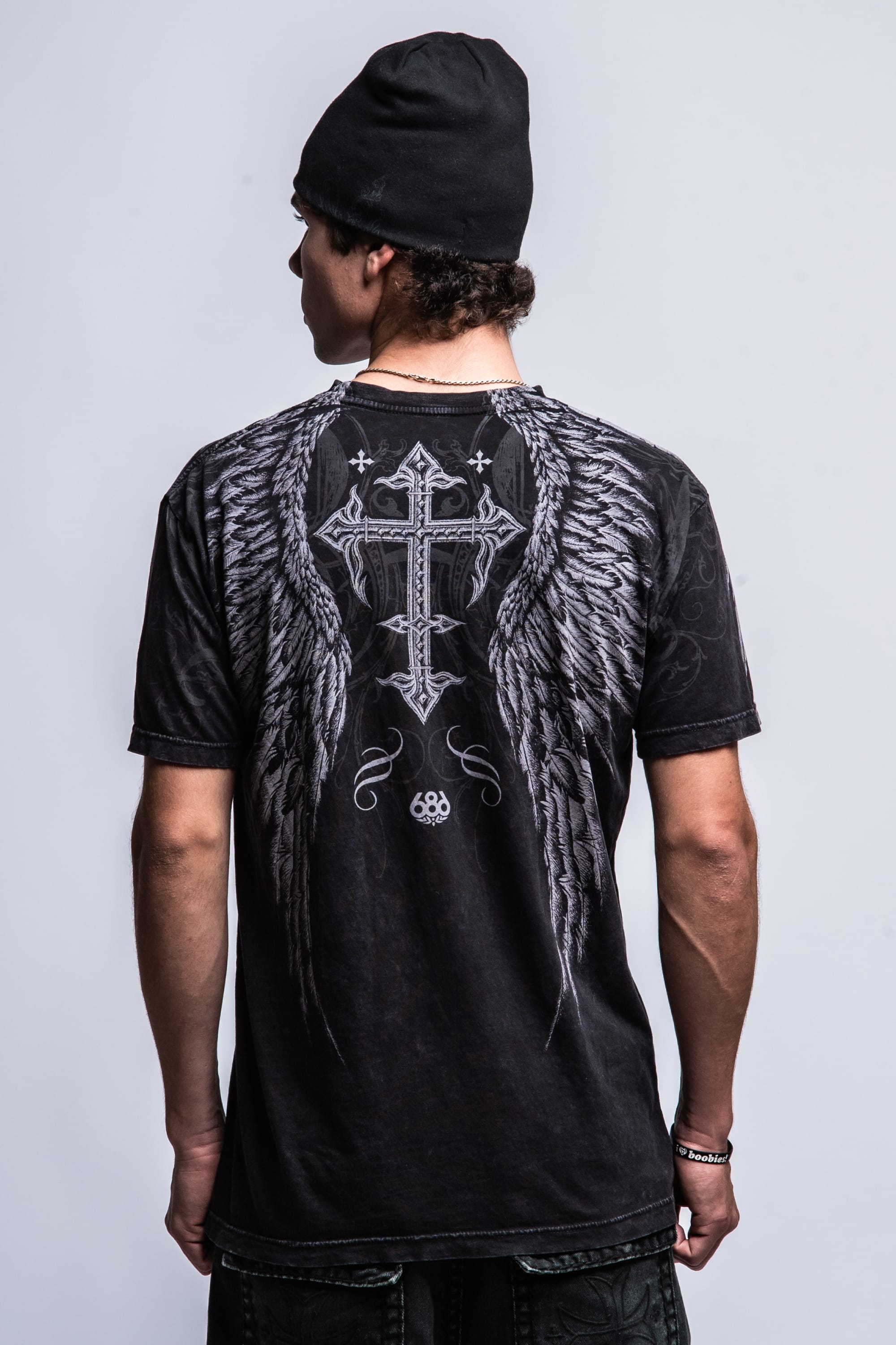A man in a black beanie and the 686 Affliction Death Eyes SS Tee by 686, featuring large angel wings and a decorative cross graphic on the back, stands against a plain light gray background.
