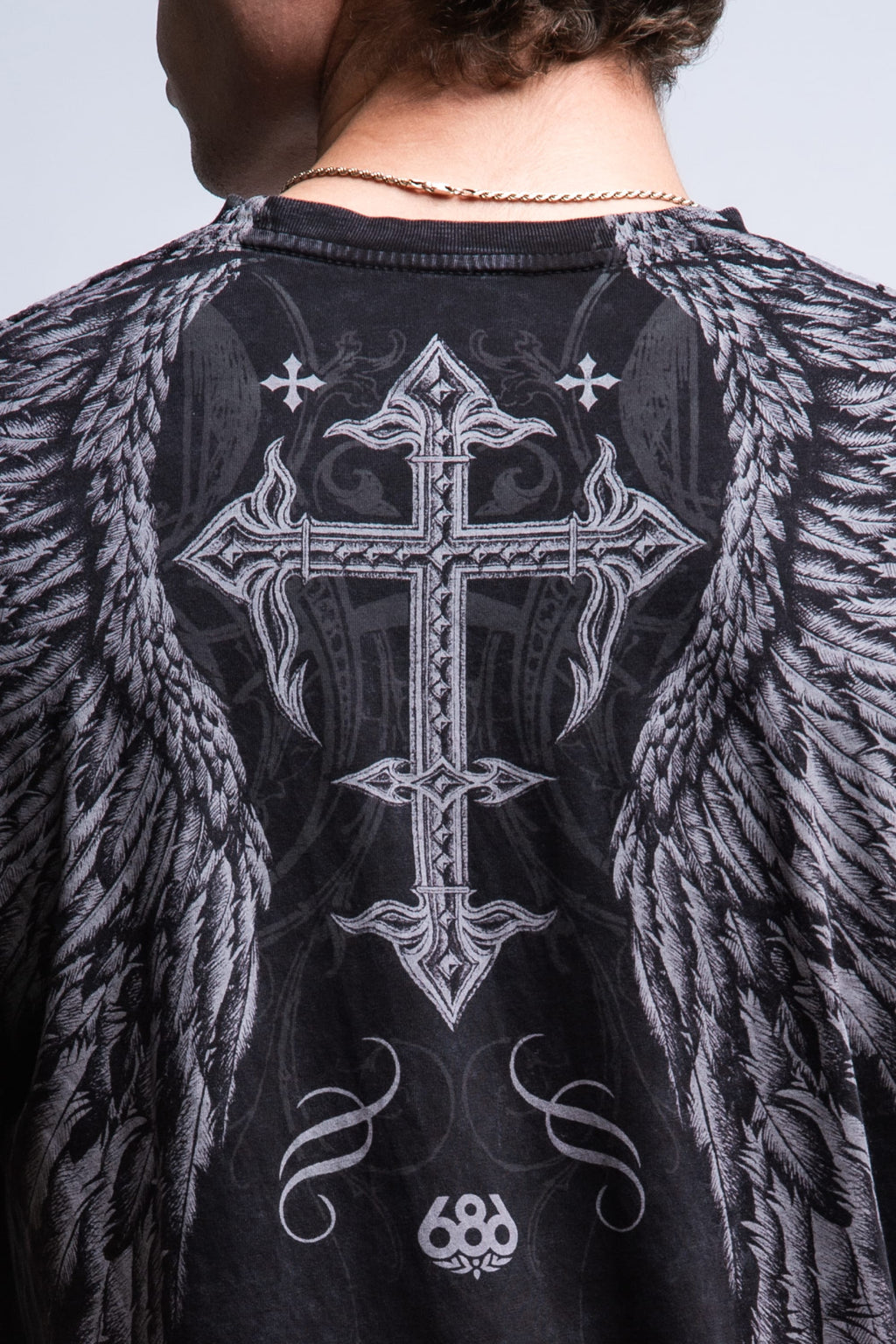 A person with short curly hair and a chain necklace wears the 686 Affliction Death Eyes SS Tee, a black 100% cotton men's t-shirt by 686 featuring a large ornate cross and angel wings design on the back with "686" near the hem.
