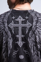 A person with short curly hair and a chain necklace wears the 686 Affliction Death Eyes SS Tee, a black 100% cotton men's t-shirt by 686 featuring a large ornate cross and angel wings design on the back with "686" near the hem.