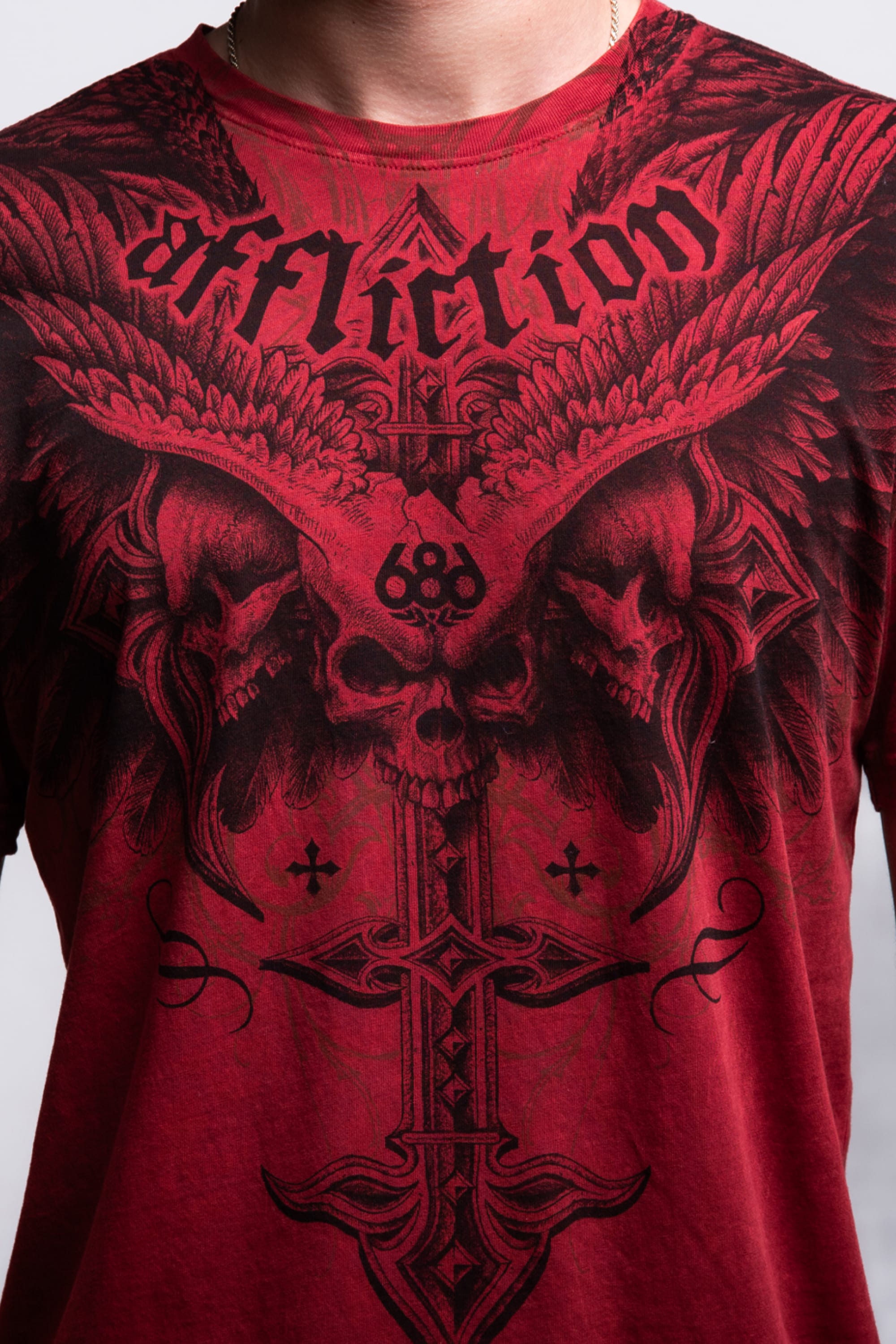 A person wearing the 686 Affliction Death Eyes SS Tee by 686, a men’s 100% cotton shirt with black gothic artwork—skulls, wings, crosses, and "Affliction" across the chest. Image shows only the chest and neck.