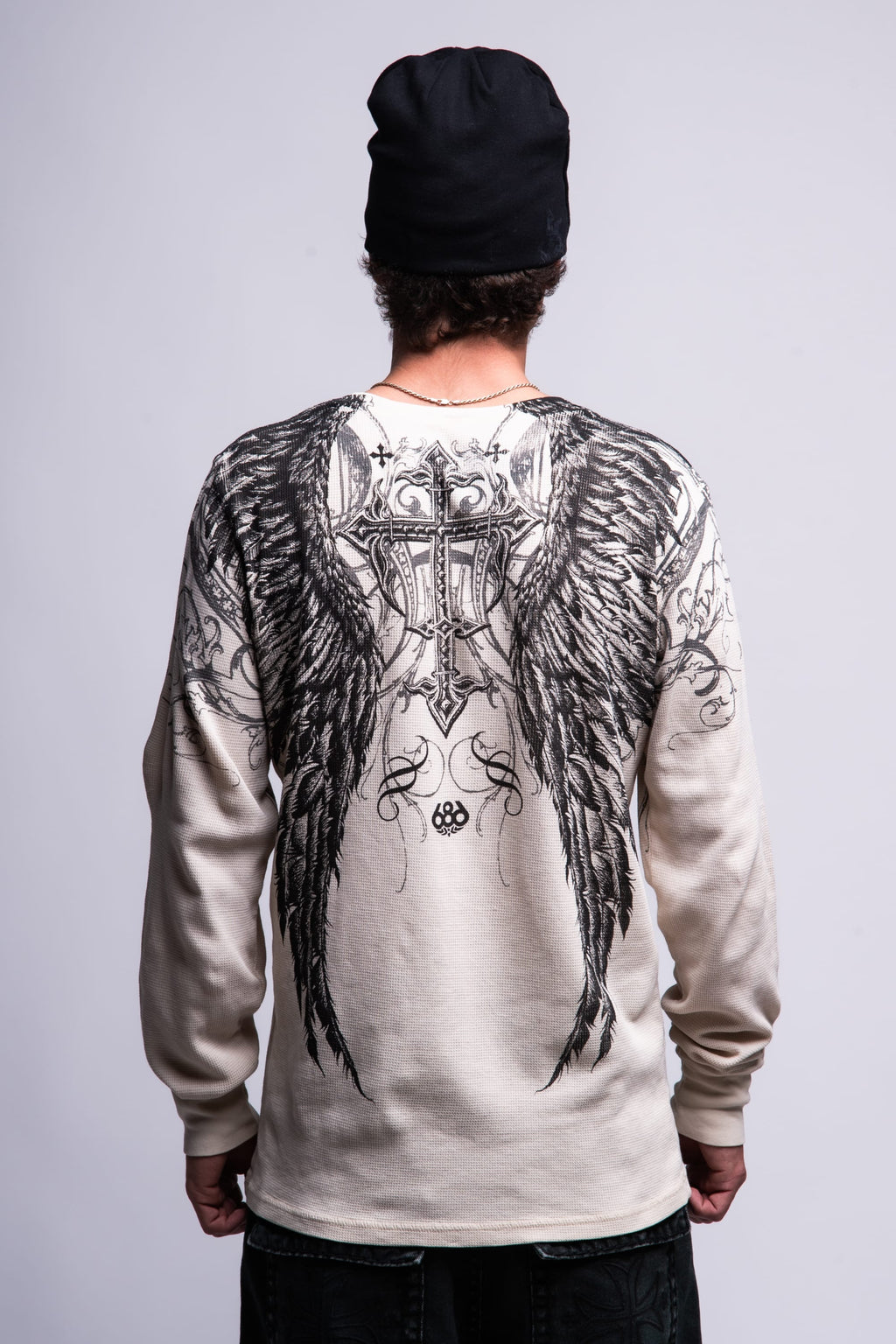 A person wearing a black beanie and the 686 Affliction Death Eyes Thermal LS by 686, a light-colored long-sleeve men's t-shirt featuring large, detailed black angel wings and a cross design on the back, stands with their back to the camera.