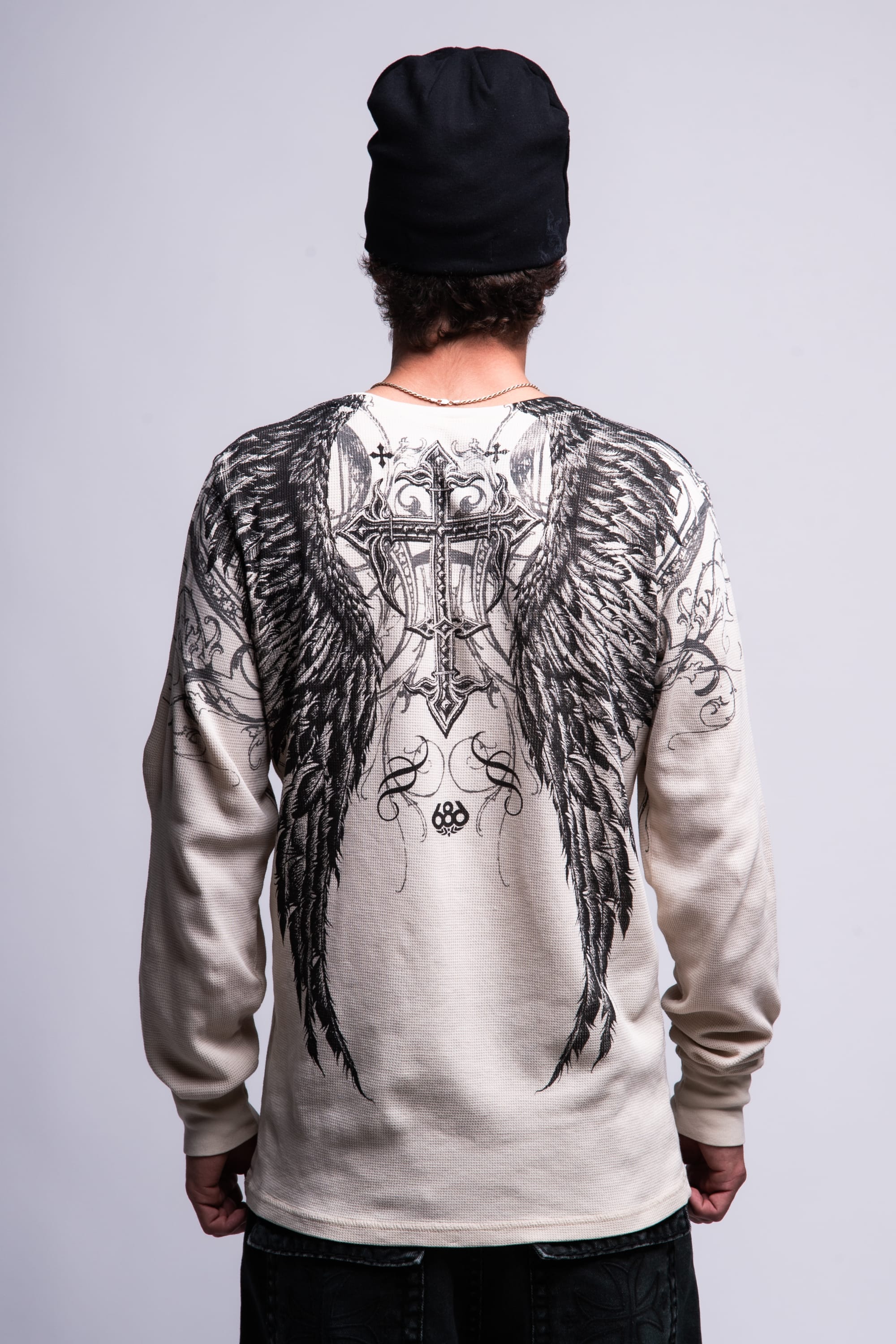 A person wearing a black beanie and the 686 Affliction Death Eyes Thermal LS by 686, a light-colored long-sleeve men's t-shirt featuring large, detailed black angel wings and a cross design on the back, stands with their back to the camera.