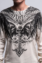 A person wears the 686 Affliction Death Eyes Thermal LS by 686—a light-colored, 100% cotton men’s tee with bold black graphics of skulls, wings, a cross, and “Affliction” in stylized text. Only their torso and part of neck are visible.