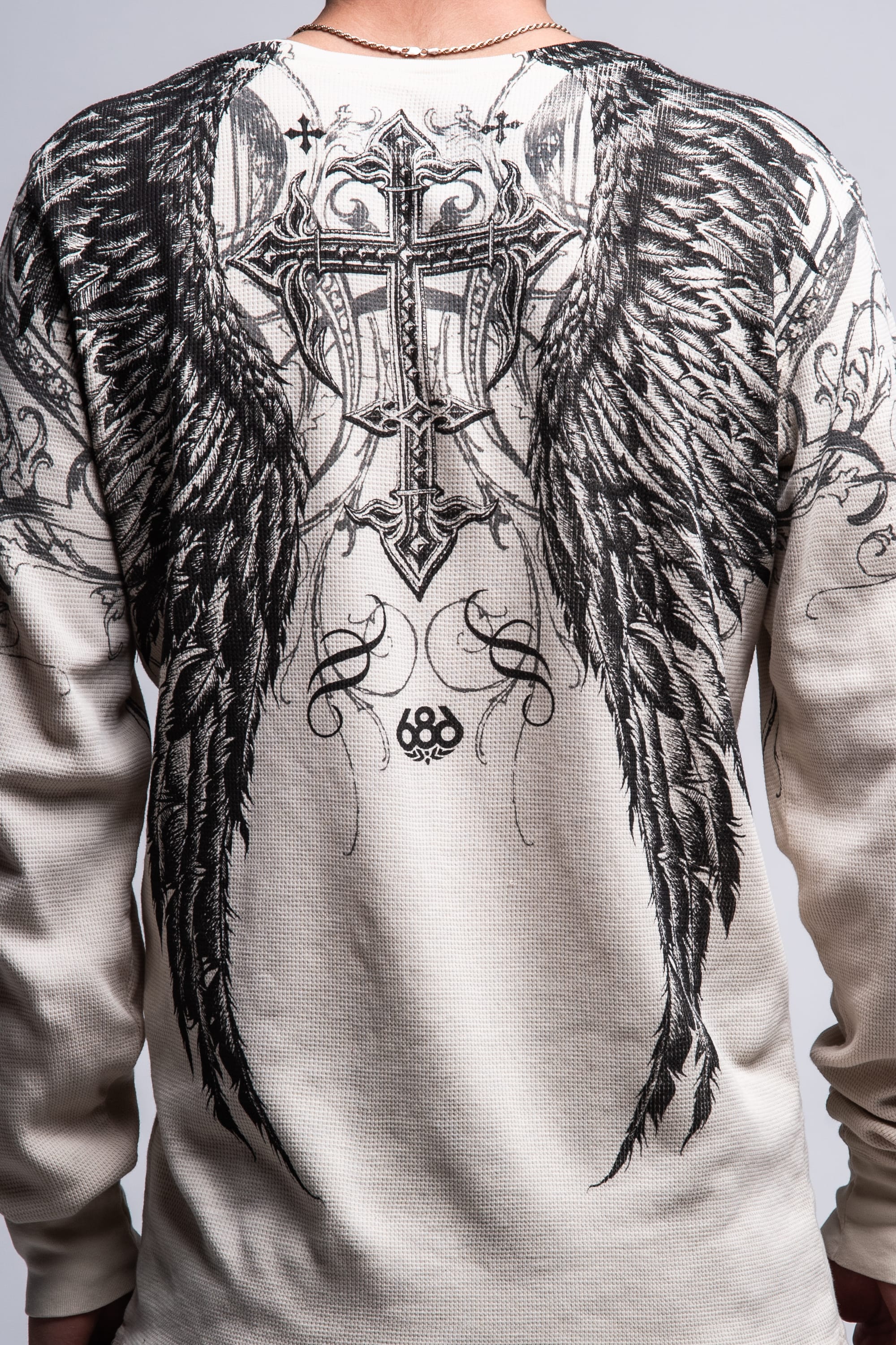 A person wears the 686 Affliction Death Eyes Thermal LS by 686, a light men's tee with black angel wings and an ornate cross graphic on the back, decorative swirls, and "686" at the center bottom. Made of soft 100% cotton for comfort.