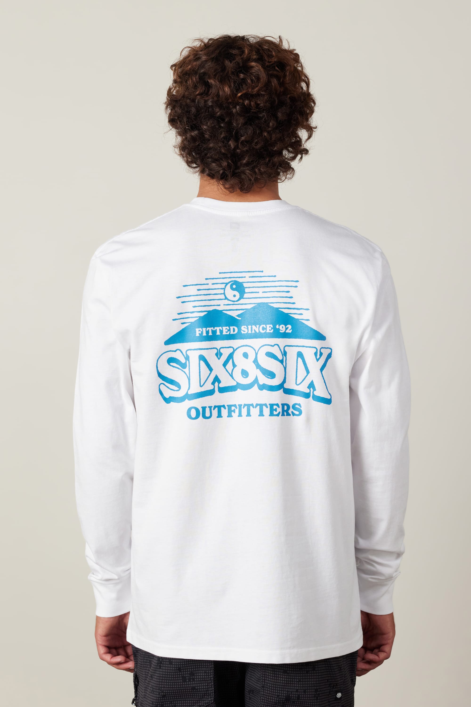 A person with curly hair, seen from behind, wears the 686 Fitted Authentic L/S Tee. This men's white long-sleeve shirt is 100% cotton and features a blue back graphic reading "Fitted Since 92," "SIX8SIX," and "OUTFITTERS.
