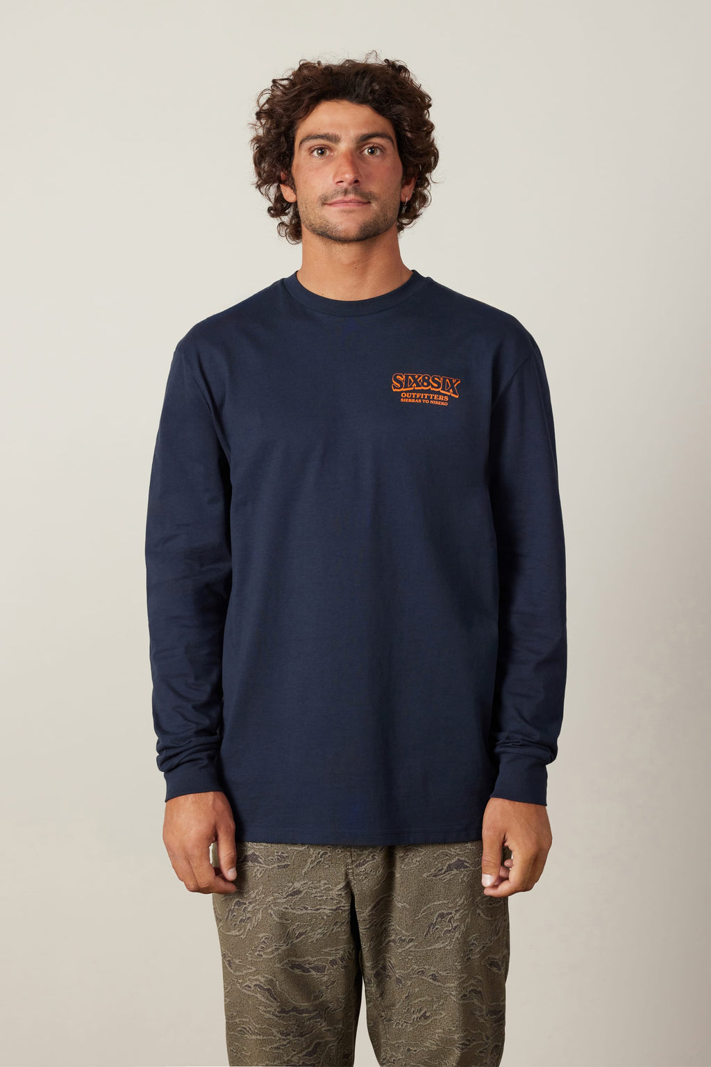 A man with curly hair wears the 686 Fitted Authentic L/S Tee by 686, a navy blue 100% cotton men’s long-sleeve shirt with orange left-chest text, paired with patterned khaki pants, standing against a plain light background.
