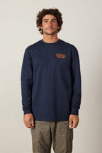 A man with curly hair wears the 686 Fitted Authentic L/S Tee by 686, a navy blue 100% cotton men’s long-sleeve shirt with orange left-chest text, paired with patterned khaki pants, standing against a plain light background.