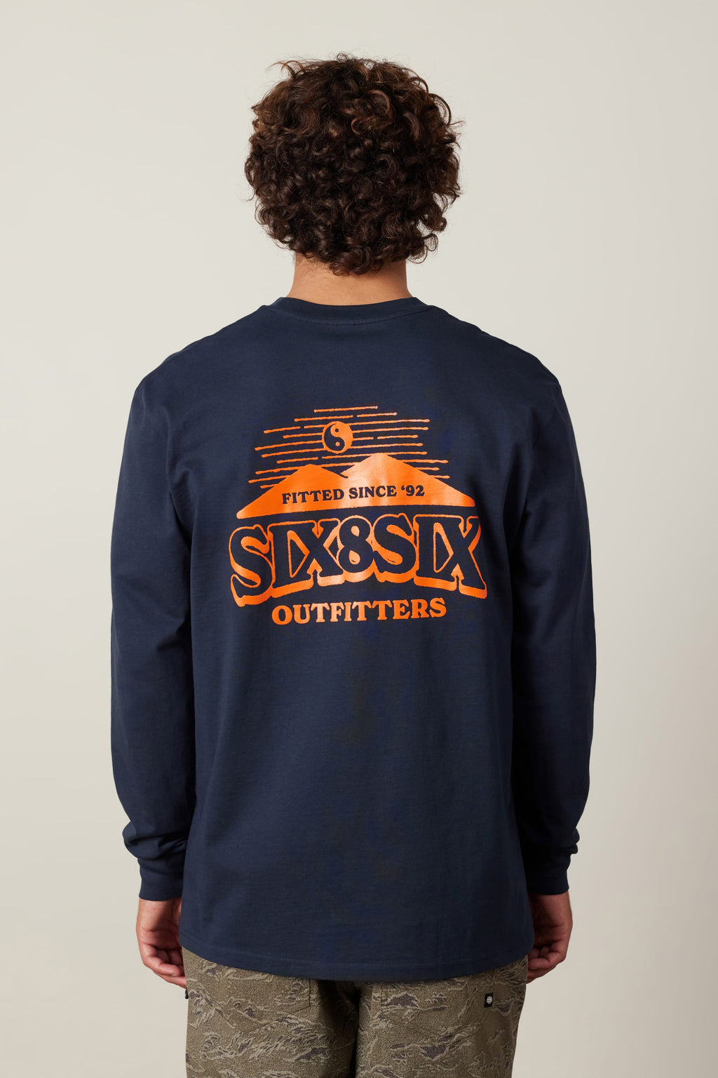 A person with curly hair, facing away, wears the navy 686 Fitted Authentic L/S Tee by 686—100% cotton with an orange “Fitted Since 92, SIX&SIX OUTFITTERS” graphic on the back.
