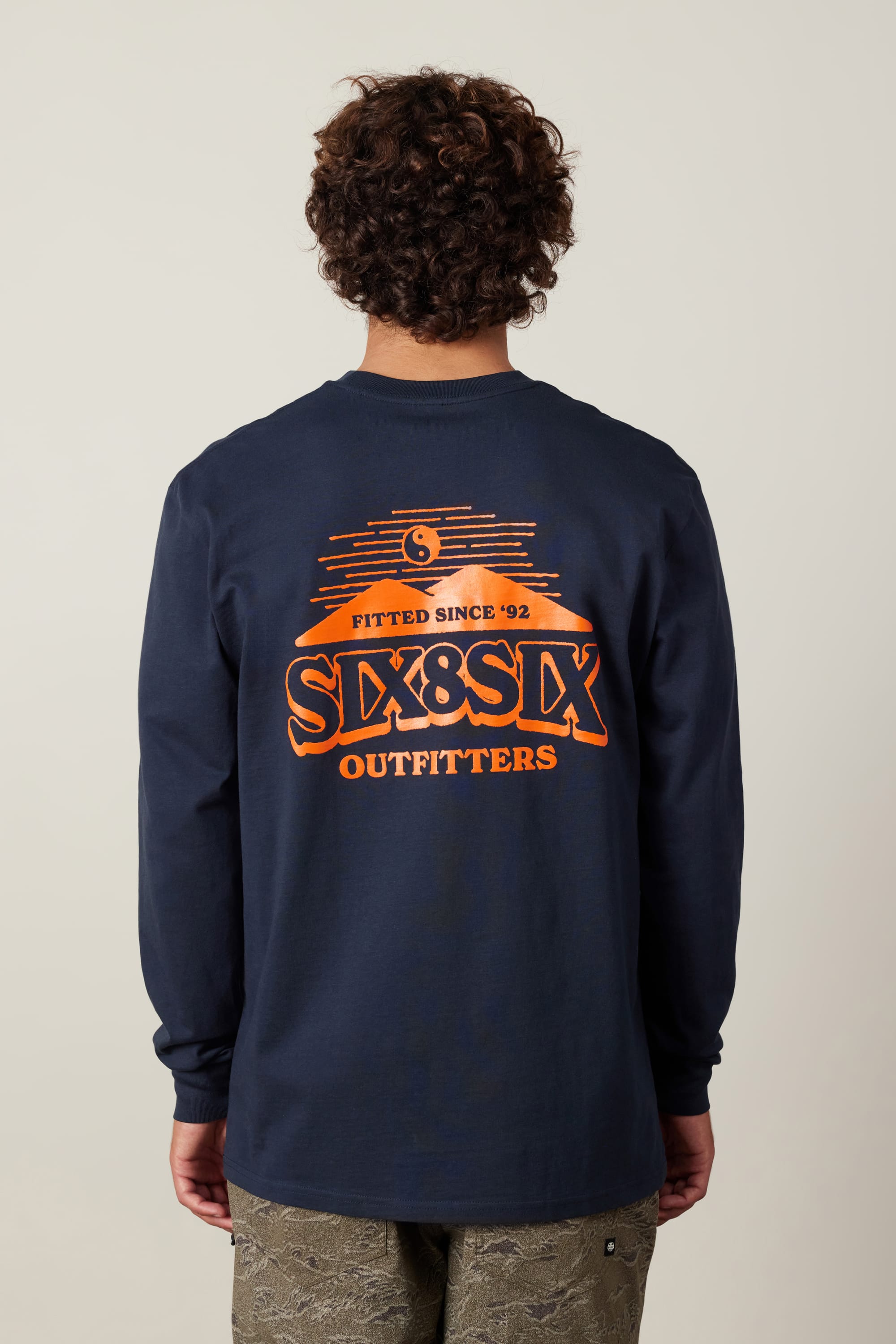 A person with curly hair, facing away, wears the navy 686 Fitted Authentic L/S Tee by 686—100% cotton with an orange “Fitted Since 92, SIX&SIX OUTFITTERS” graphic on the back.
