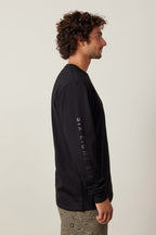 A man with curly hair stands in profile wearing the 686 High Def Authentic L/S Tee by 686, featuring "SIX EIGHT SIX" on the sleeve. His light patterned pants contrast the 100% cotton shirt, set against a plain light background.