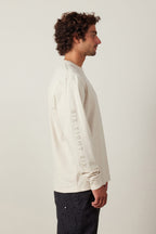 A man with curly hair stands in profile, wearing a 686 High Def Authentic L/S Tee in light beige, made of 100% cotton with “SIX EIGHT SIX” on the sleeve, paired with dark patterned pants against a plain light background.