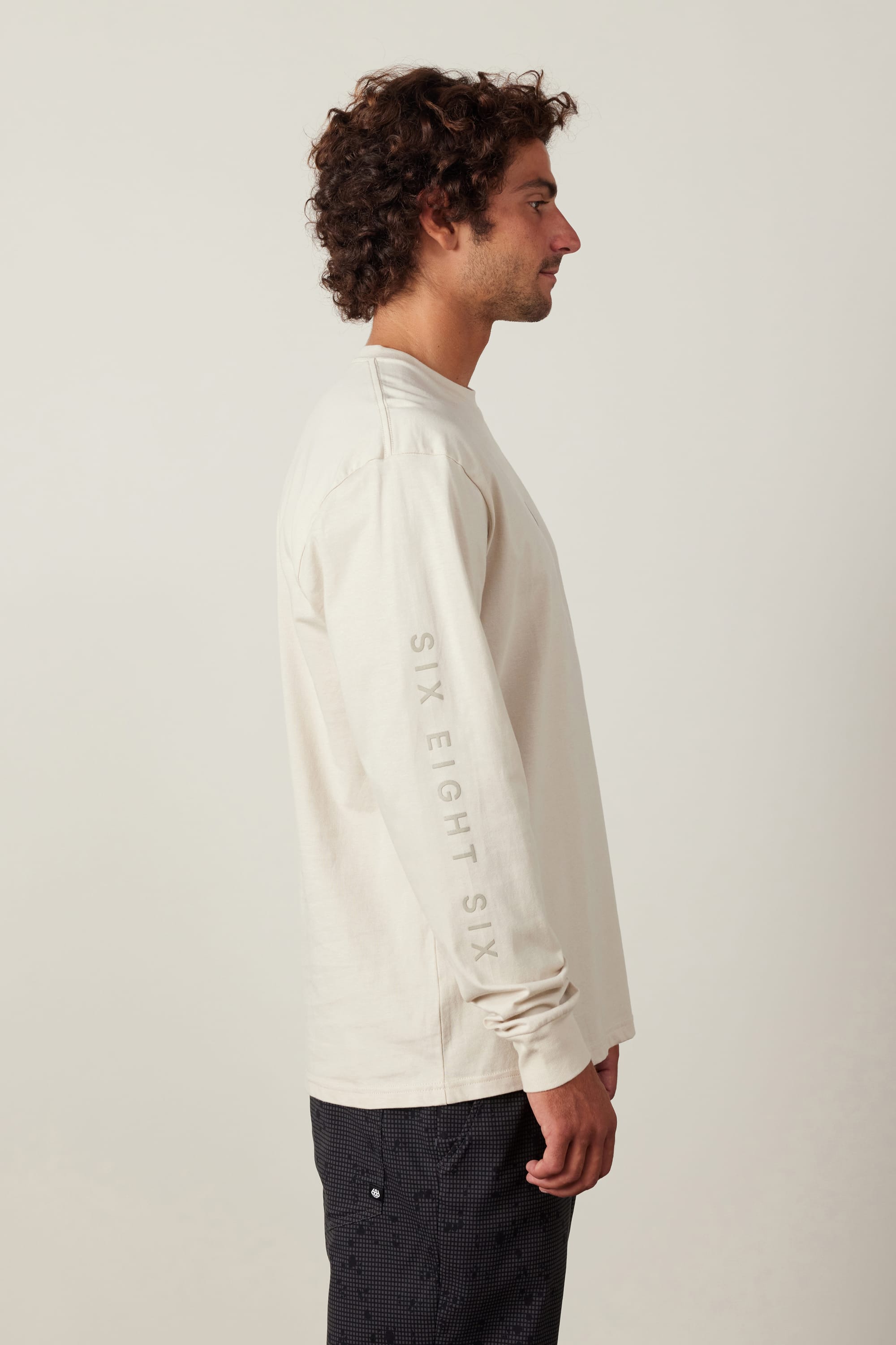 A man with curly hair stands in profile, wearing a 686 High Def Authentic L/S Tee in light beige, made of 100% cotton with “SIX EIGHT SIX” on the sleeve, paired with dark patterned pants against a plain light background.
