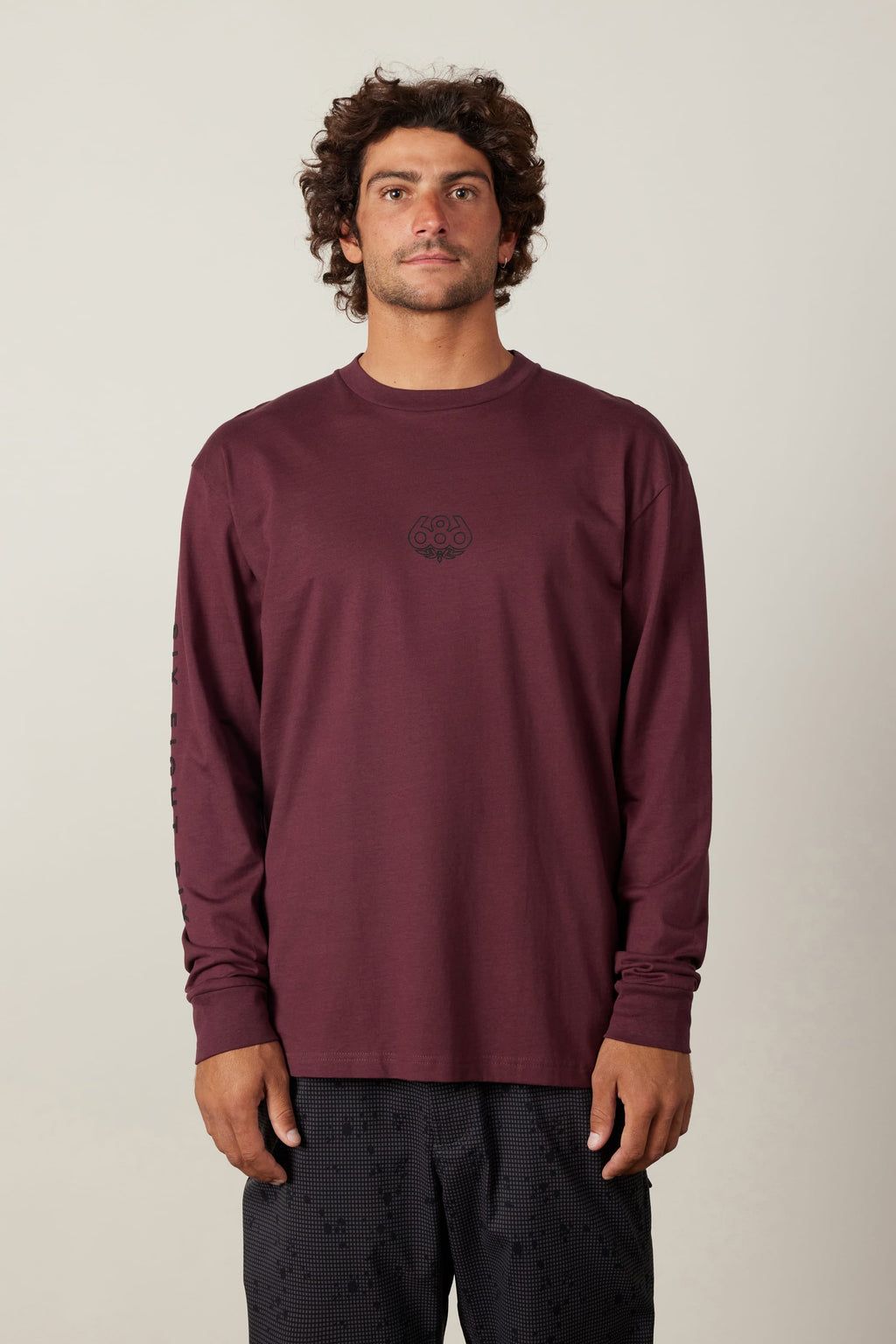 A man with curly hair wears the 686 High Def Authentic L/S Tee, a burgundy long-sleeve shirt with a small black chest logo by 686, paired with black patterned shorts, standing against a plain light background.