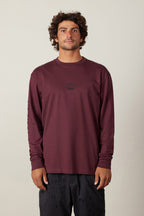 A man with curly hair wears the 686 High Def Authentic L/S Tee, a burgundy long-sleeve shirt with a small black chest logo by 686, paired with black patterned shorts, standing against a plain light background.