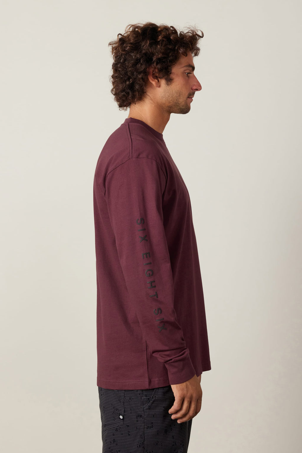 A man with curly hair stands sideways against a plain light background, wearing the 686 High Def Authentic L/S Tee by 686 and dark patterned pants.