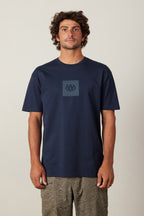 A man with curly hair faces forward wearing the 686 Knockout Authentic S/S Tee in navy blue, featuring a small 686 logo on the chest, paired with olive green patterned shorts. The background is plain and light-colored.