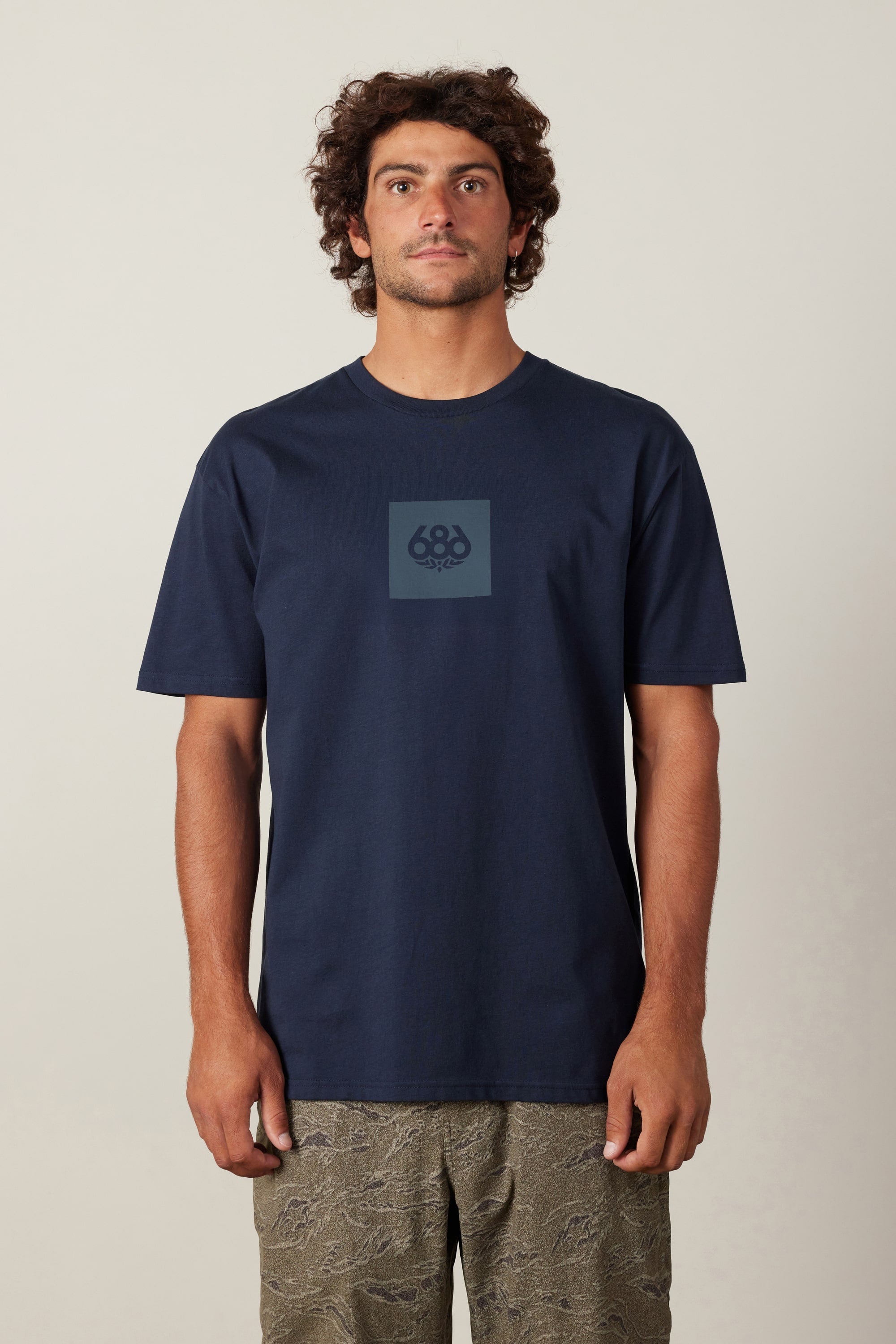 A man with curly hair faces forward wearing the 686 Knockout Authentic S/S Tee in navy blue, featuring a small 686 logo on the chest, paired with olive green patterned shorts. The background is plain and light-colored.