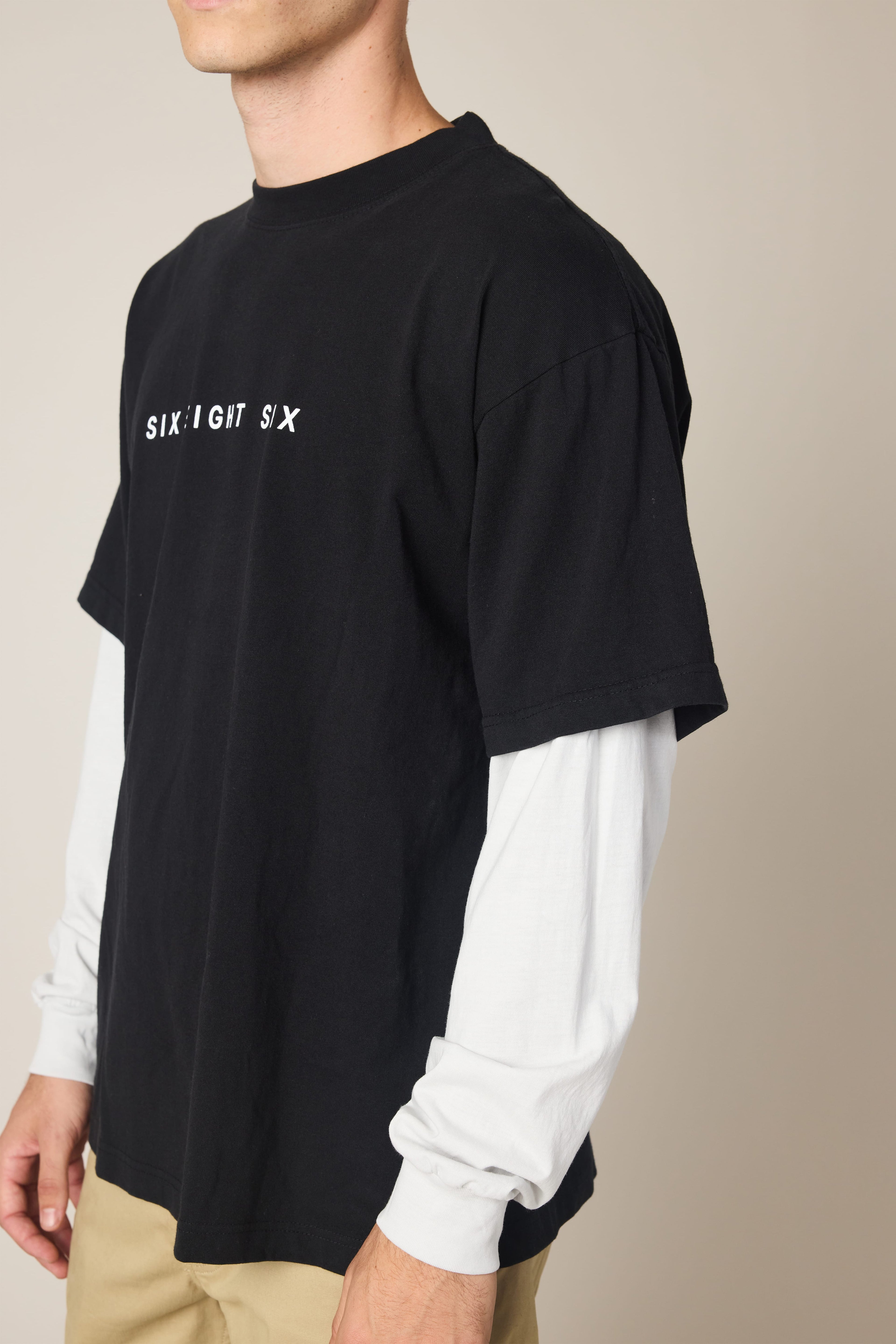 A person wearing the 686 Wordmark Relaxed 2fer LS Tee—a black men's cotton t-shirt with "SIX:EIGHT SIX" in white over long white sleeves—with beige pants stands against a plain background, their face partially out of frame.