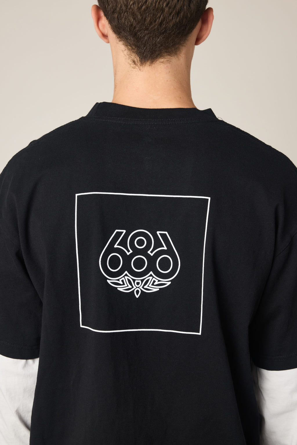 A person shown from behind wears the 686 Wordmark Relaxed 2fer LS Tee, a black 100% cotton men’s t-shirt by 686, featuring a white outlined square with the 686 logo and graphic design on the back, complementing their short brown hair.
