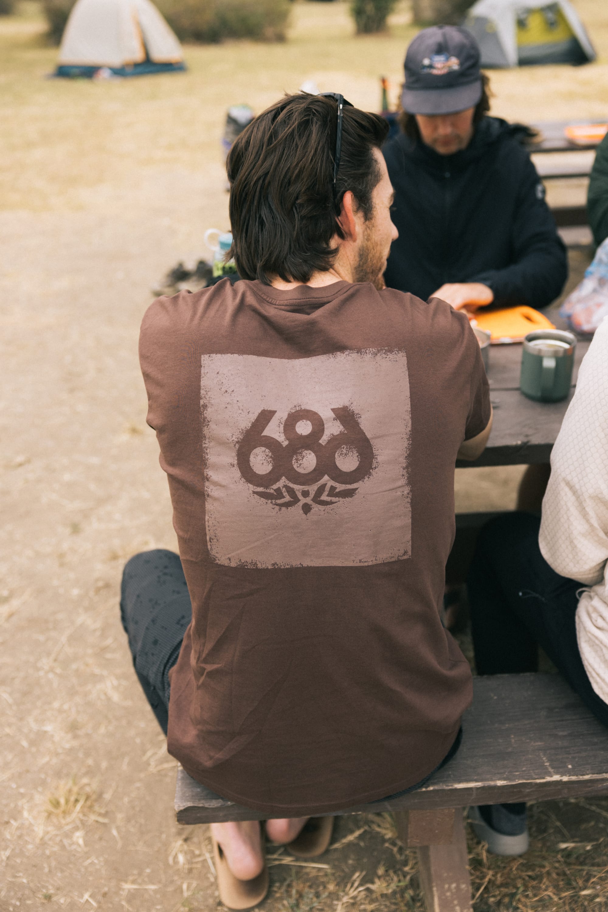 A man with dark hair sits at an outdoor picnic table wearing the 686 Knockout Imprint Authentic S/S Tee by 686, which features a large graphic on the back. Other people and tents in the background suggest a camping scene.