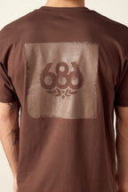 A person wears the 686 Knockout Imprint Authentic S/S Tee by 686, a brown 100% cotton mens T-shirt with a faded rectangular 686 graphic and small design on the back, paired with visible tattoos on their left arm.